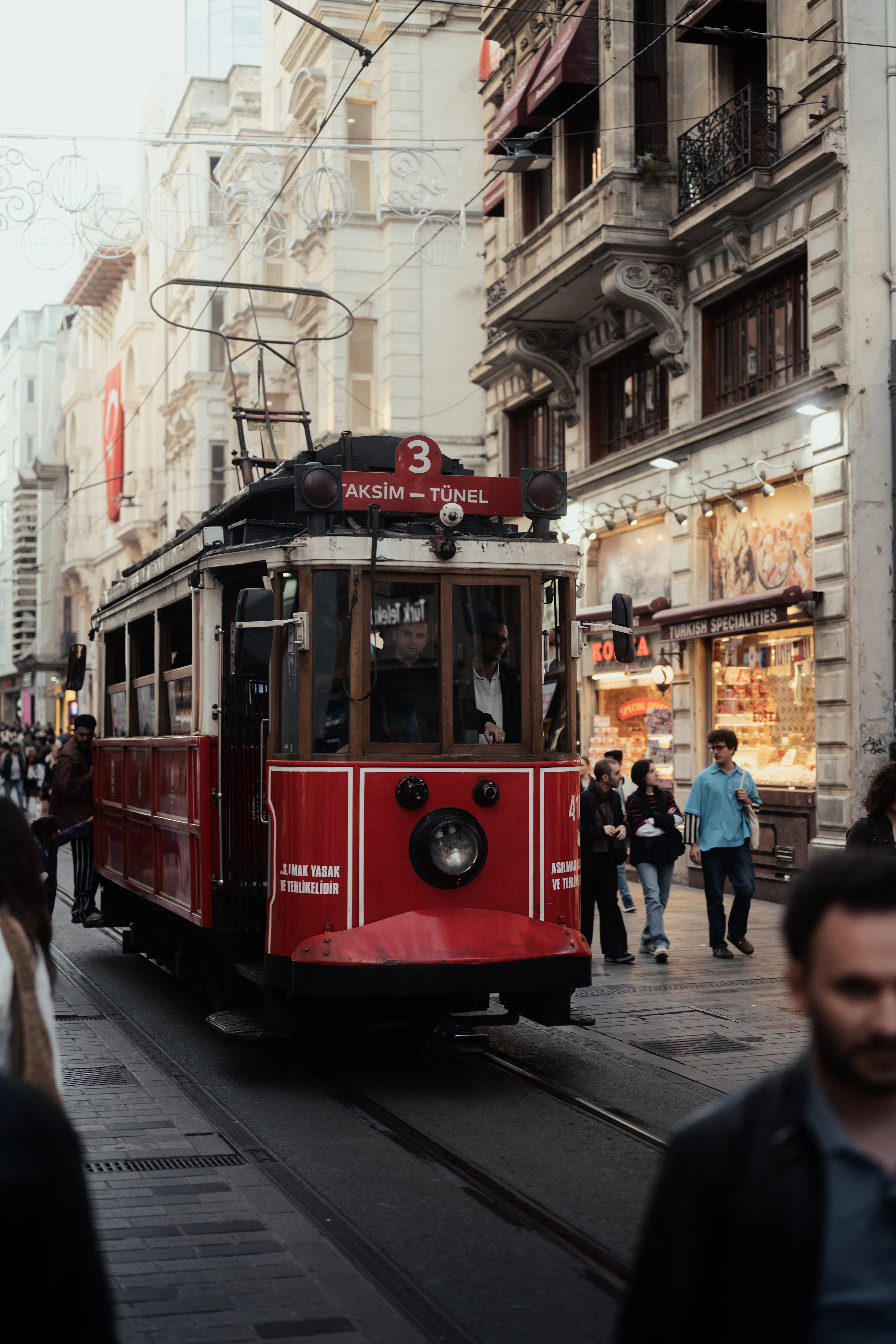 The red nostalgic tram marked Taksim-Tunel running down the middle of Istiklal Avenue in Istanbul's Beyoglu, shoppers on the pavement beside a lit shopfront and 19th-century facades overhead