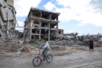Boy rides a bicycle through a war-torn area.