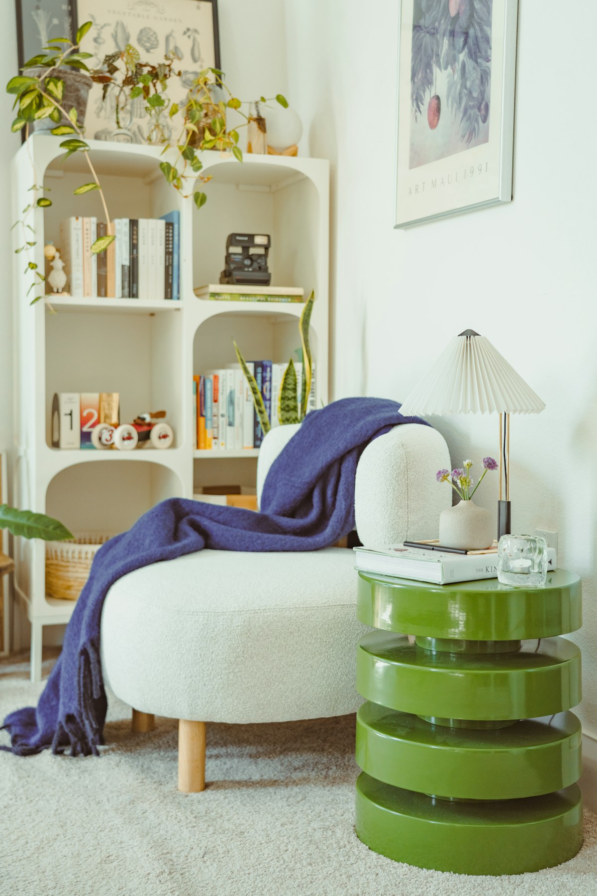 Rincón de lectura en casa con sillón blanco tapizado, manta azul, biblioteca blanca llena de libros y plantas, y una lámpara de mesa con luz cálida