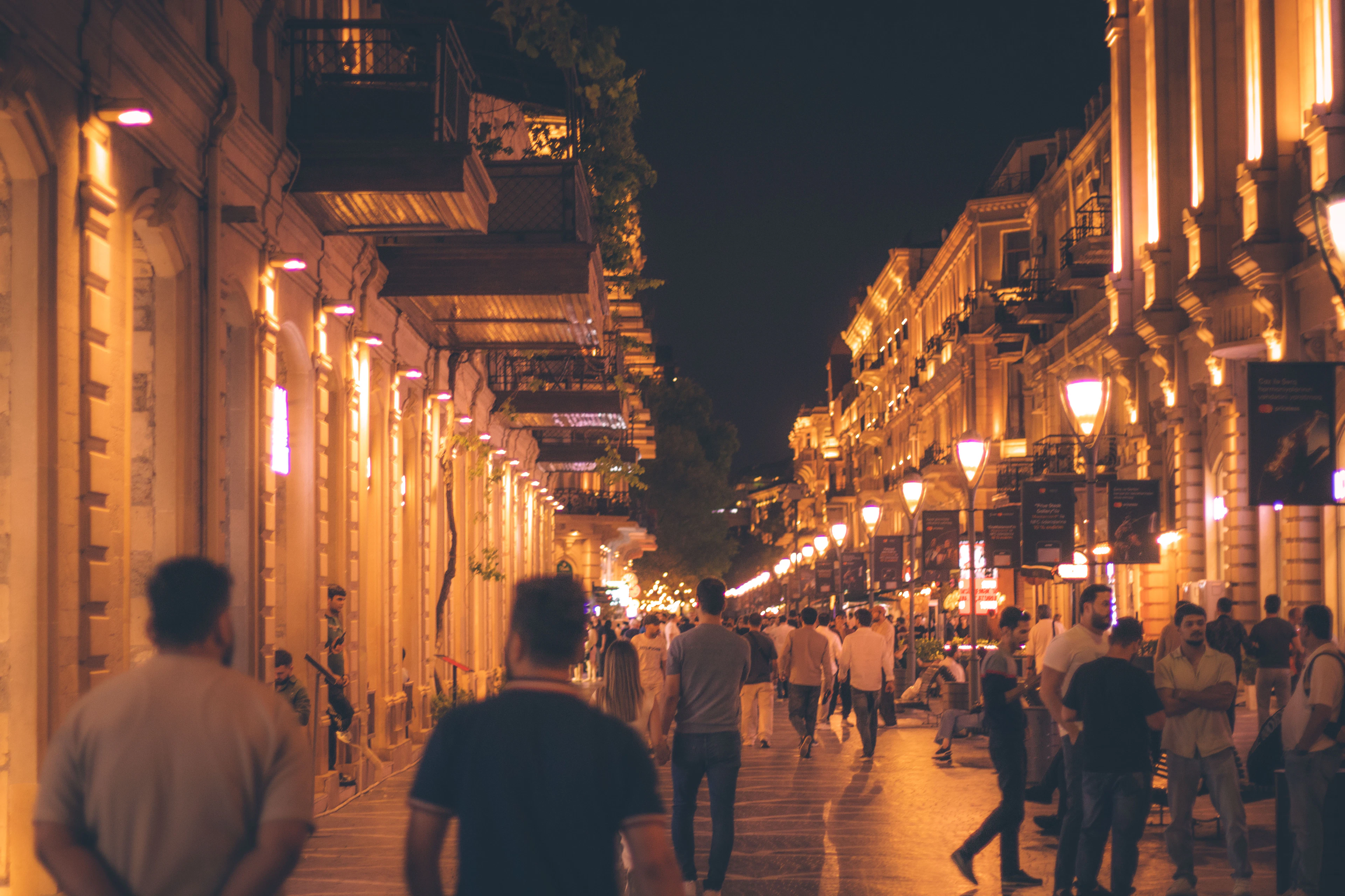 Crowds stroll a lantern-lit Baku street at night, its balconied facades glowing gold.