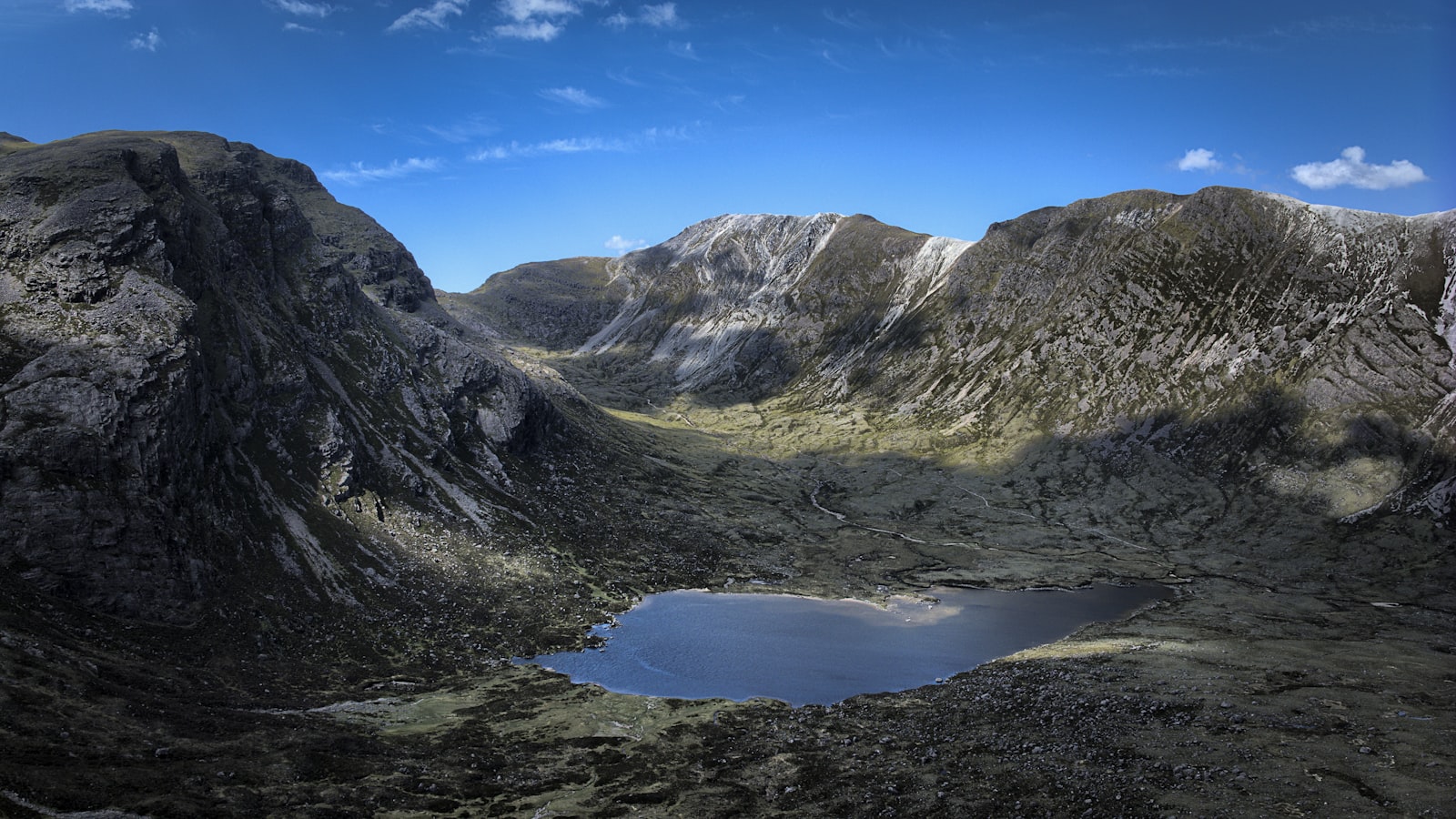 A dark lochan cupped in a wide rocky corrie ringed by steep gray-and-white banded mountainsides under a deep blue sky