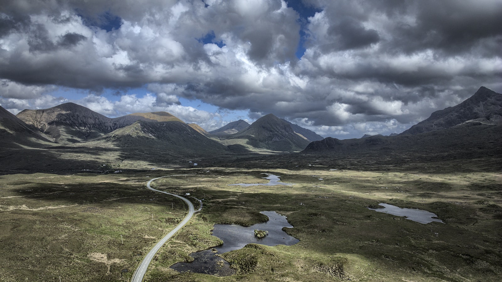 Aerial view of a single-track road winding across boggy green moorland dotted with lochans toward a range of dark, jagged mountains under heavy clouds