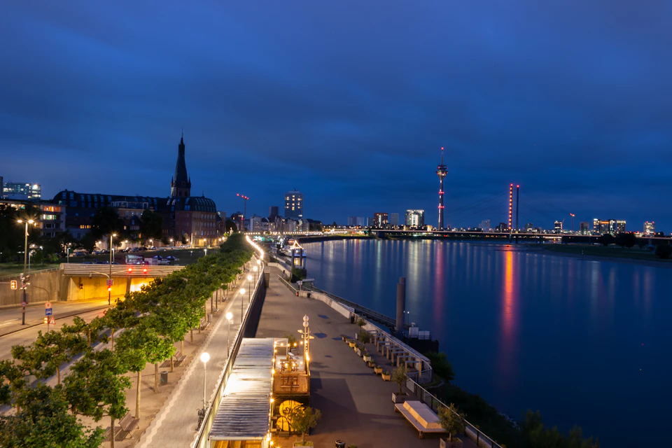 The image depicts a serene night scene along the Rhine River in Germany, with the city skyline visible in the background. The river is illuminated by the warm glow of streetlights and the reflection of buildings on its surface. On one side of the river, there's a park filled with trees and benches, providing a peaceful spot for relaxation.

In the foreground, several boats are docked along the riverbank, adding to the picturesque view. The city skyline is visible in the background, showcasing the blend of nature and urban development.