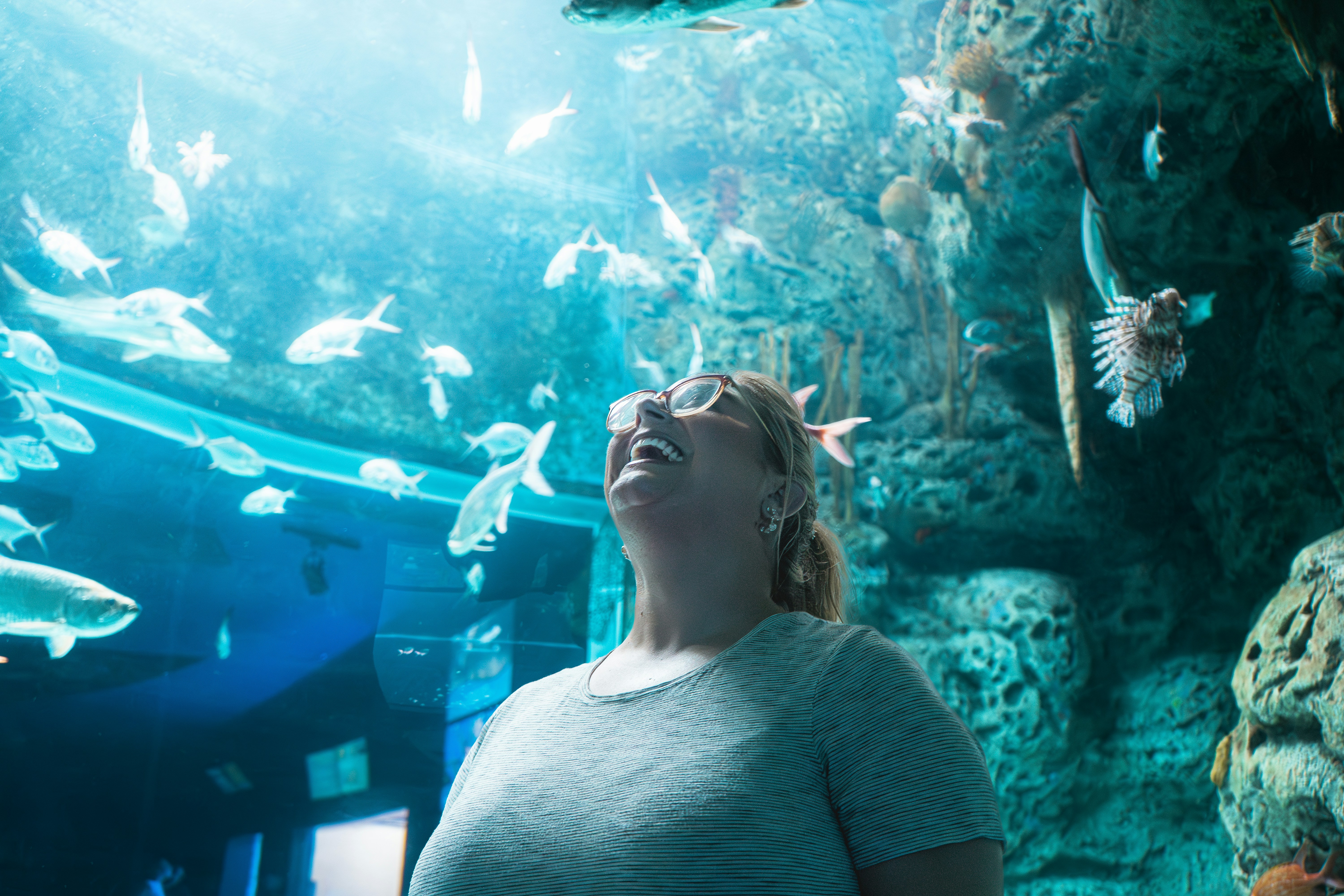 A woman with glasses looks up with a wide smile and joyful expression, completely captivated by the fish swimming in a vibrant aquarium above her. The blue water and diverse marine life create an immersive and delightful experience.