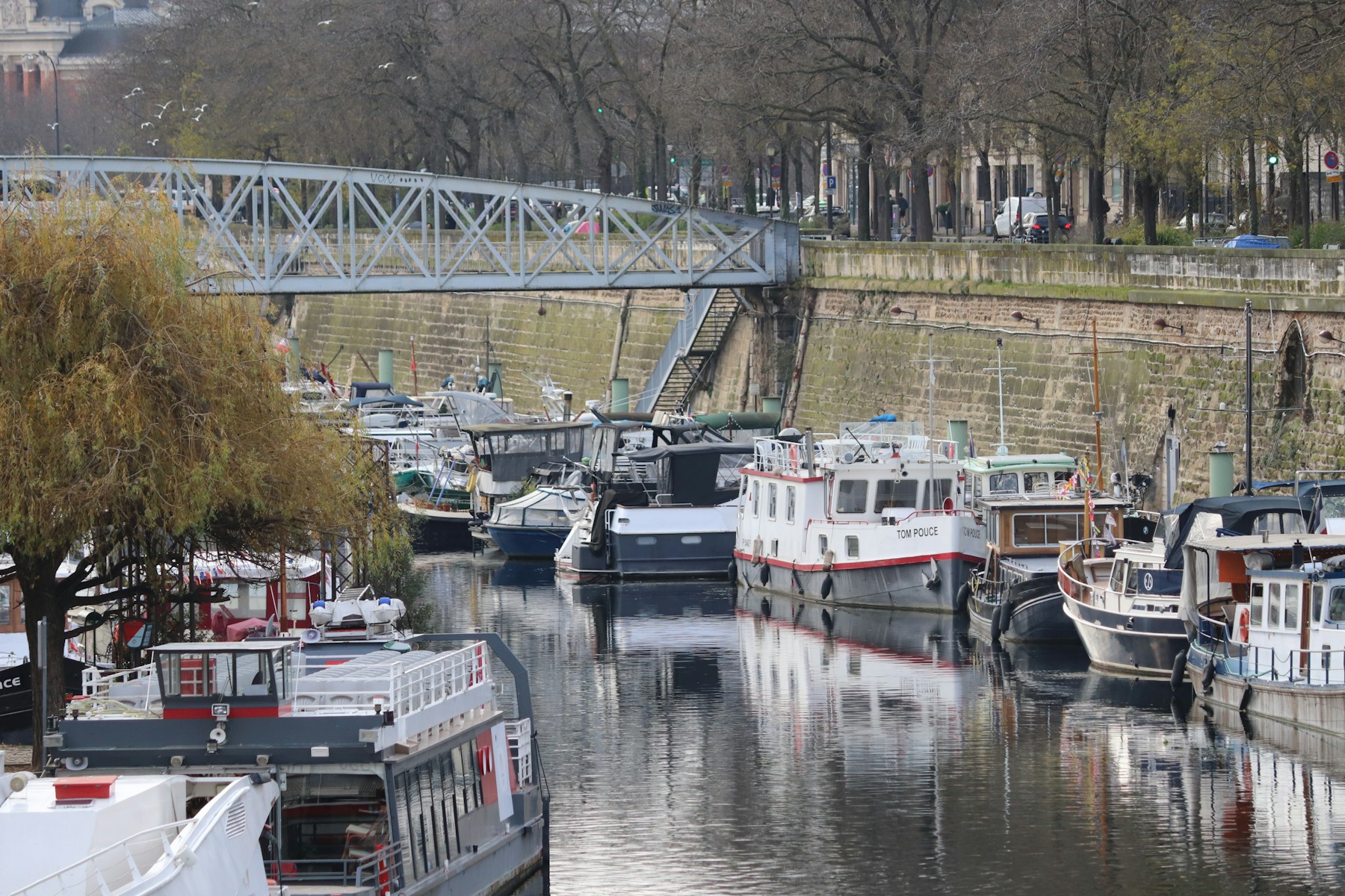 Moored boats on a tree-lined Paris canal beneath a blue pedestrian bridge.