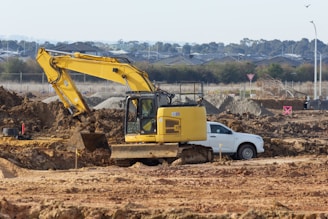 A yellow excavator is digging near a white car.