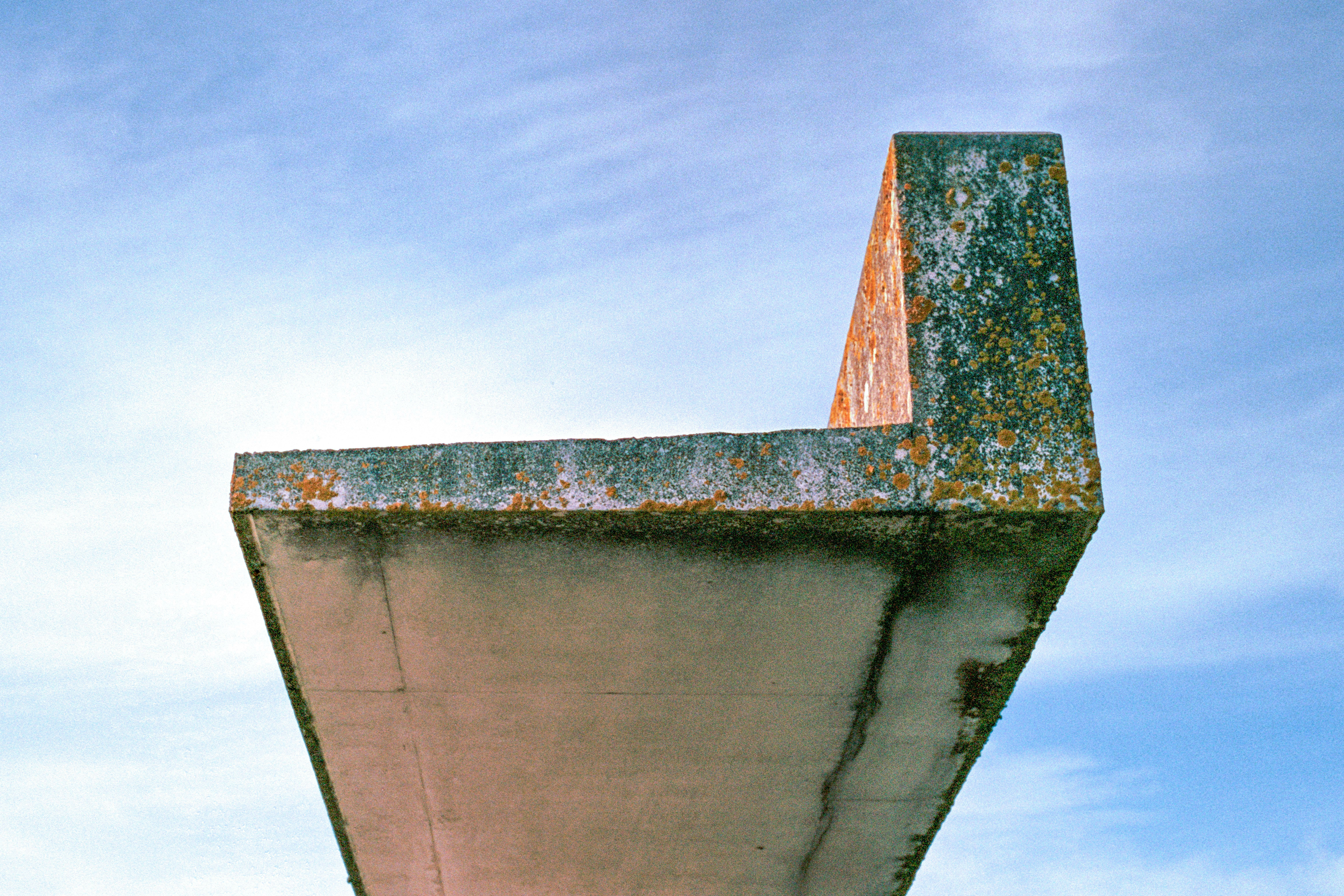Close-up view of a weathered concrete structure from the submarine base in Saint-Nazaire, France. Covered in lichen and rust stains, the structure stands out against a pale blue sky. Captured on Kodak Portra 400 film with a Nikon F3 HP.