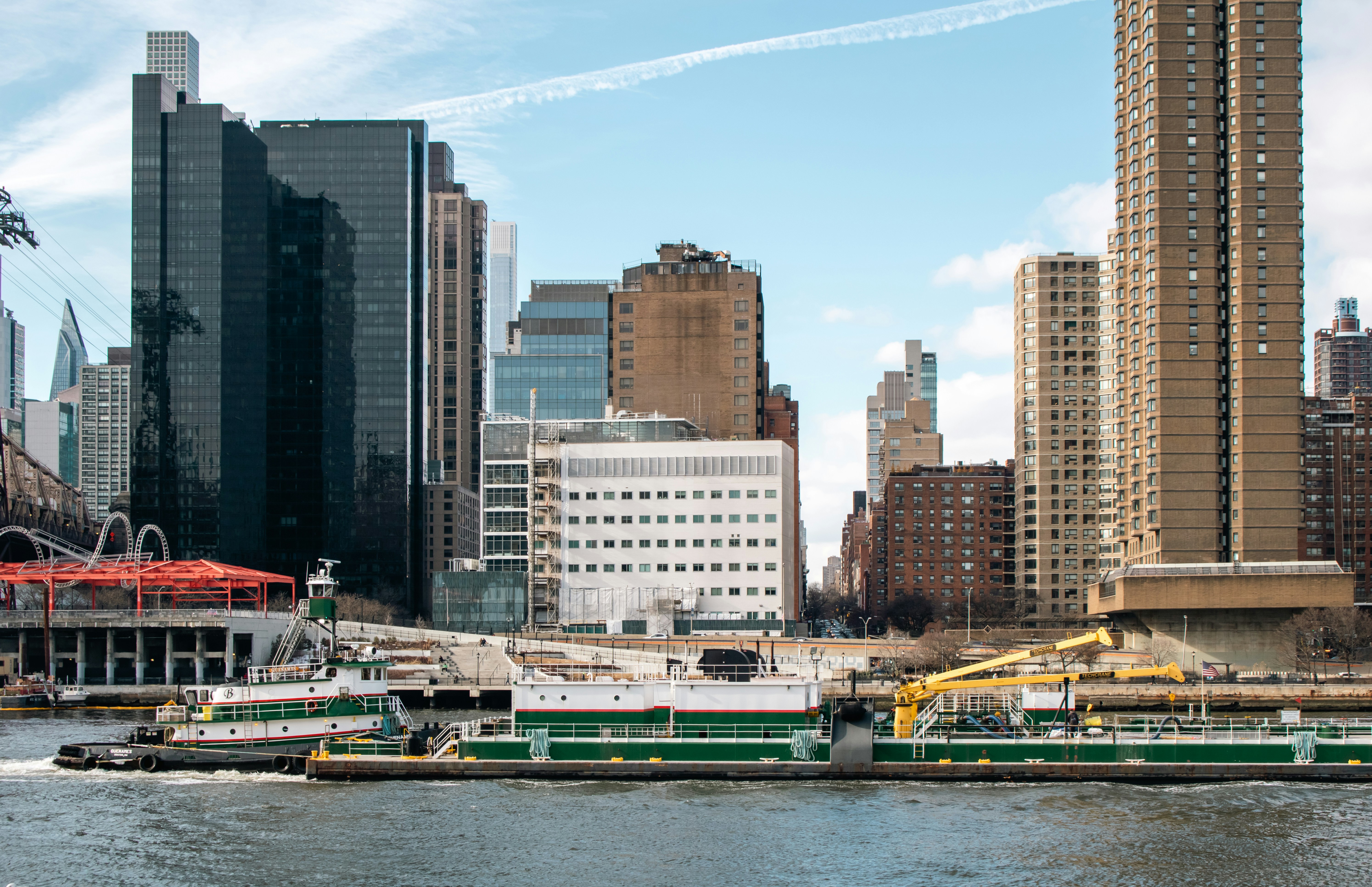 A green and white industrial barge moves along the East River, passing the East Side of Manhattan as seen from Roosevelt Island. Framed by the towering midtown buildings and the Queensboro Bridge’s edge, the vessel contrasts sharply with the city’s glassy high-rises and brick apartment blocks. This image captures the active working side of New York City’s waterways where commerce and construction quietly coexist with the ever-evolving urban skyline.