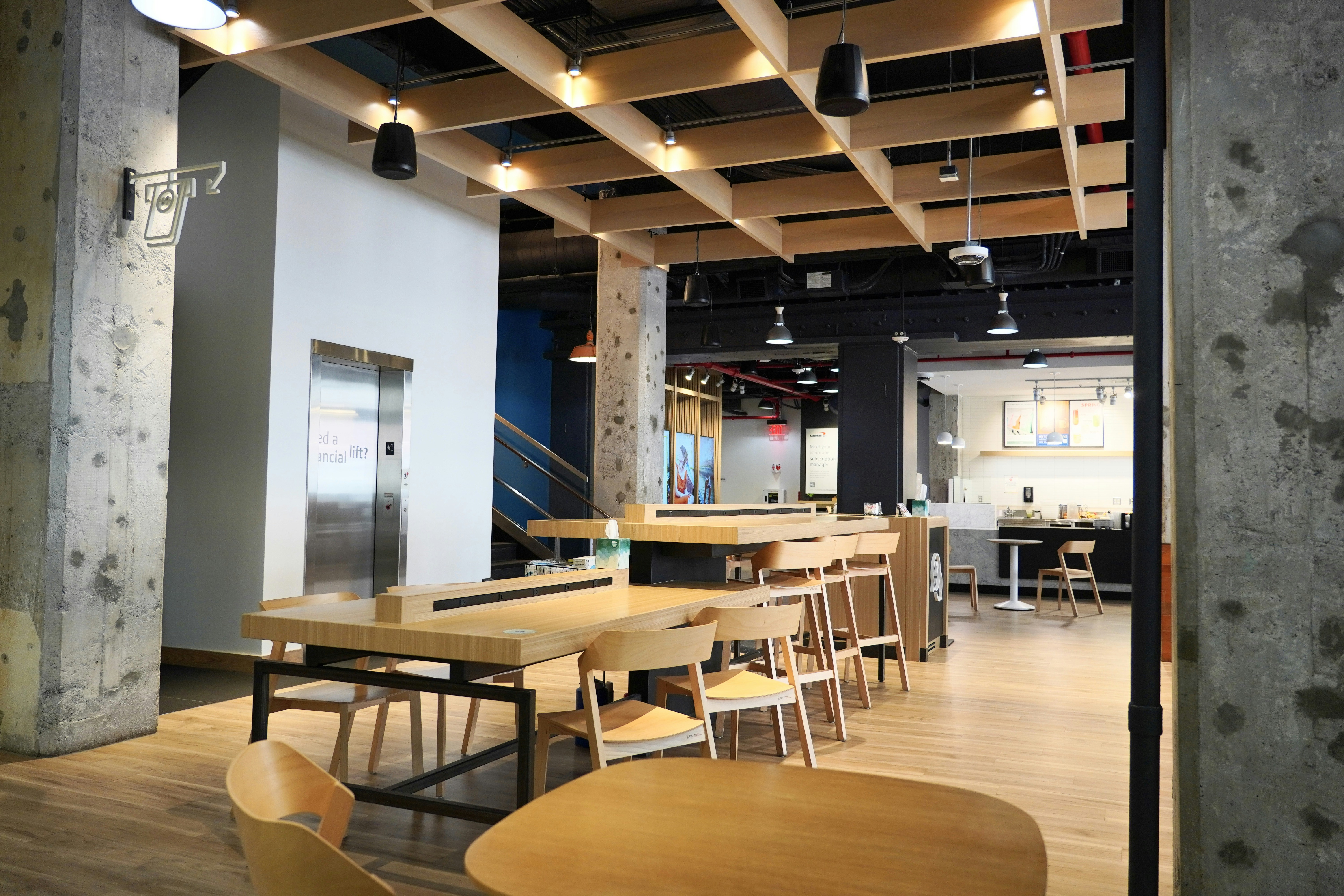 This bright, modern Capital One Café features blonde wood tables, concrete pillars, and a ceiling grid of warm lighting. Sleek chairs and communal seating reflect the hybrid function of the space, offering a relaxed spot for coffee, coworking, or financial guidance. The open-plan layout gives it a polished, minimal vibe with a welcoming feel.