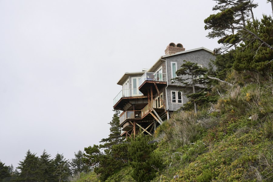 Grey shingled house with stacked decks standing on a fir-covered bluff under a soft overcast sky