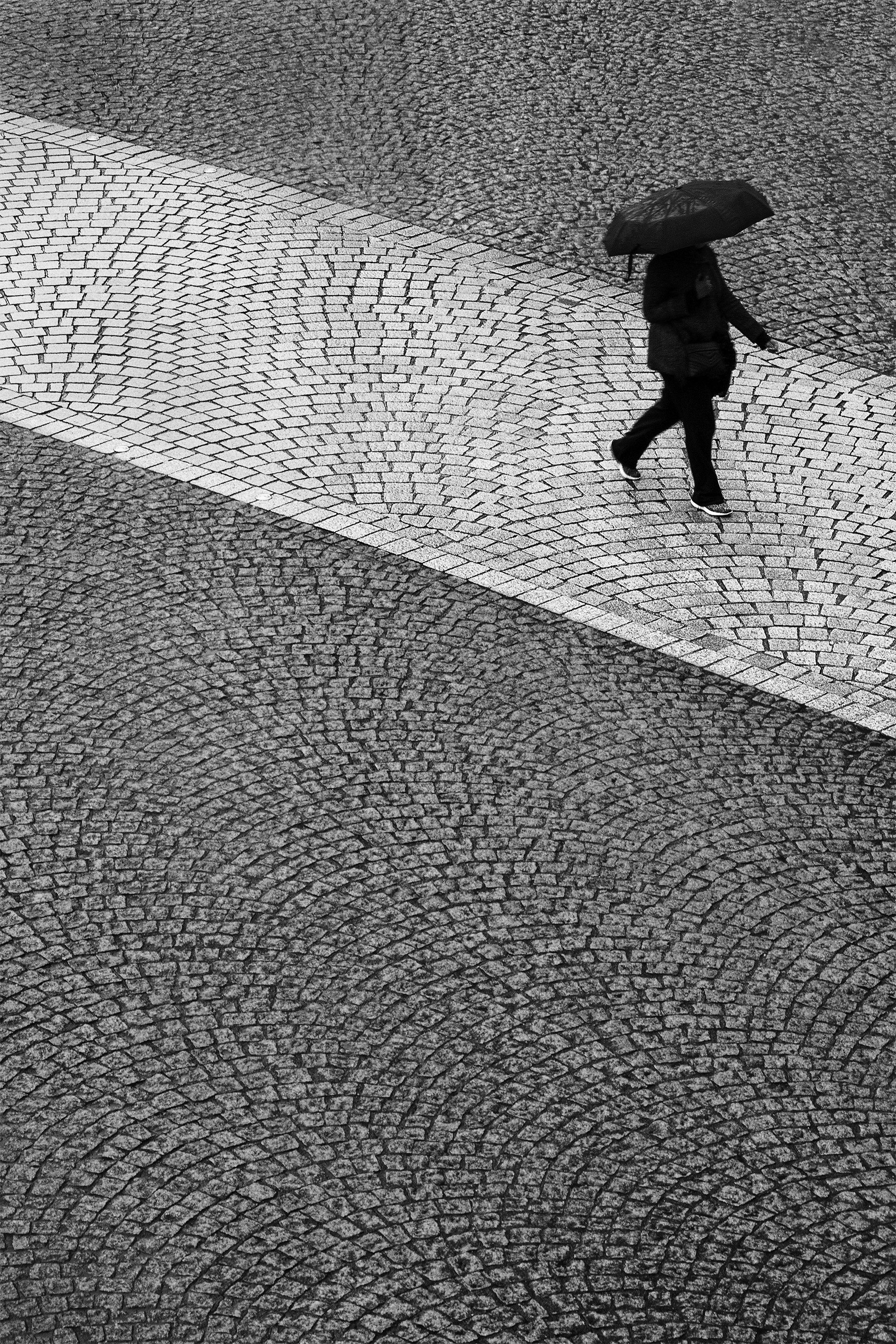Person geht mit Regenschirm auf einem Kopfsteinpflasterweg.