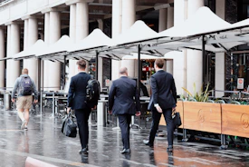 Businessmen walk down a wet street past an outdoor cafe.