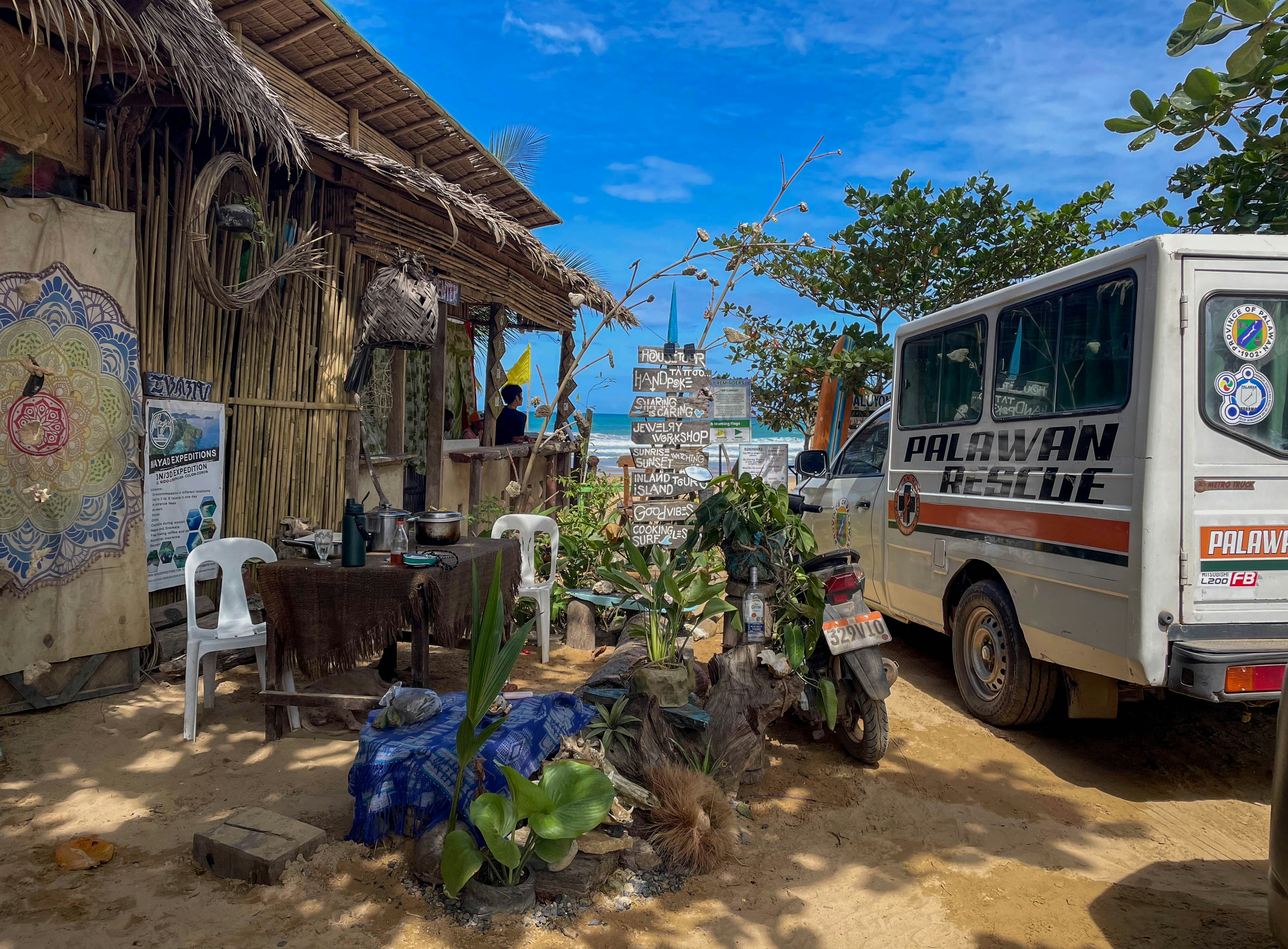 A tropical beach shack sits by a van.