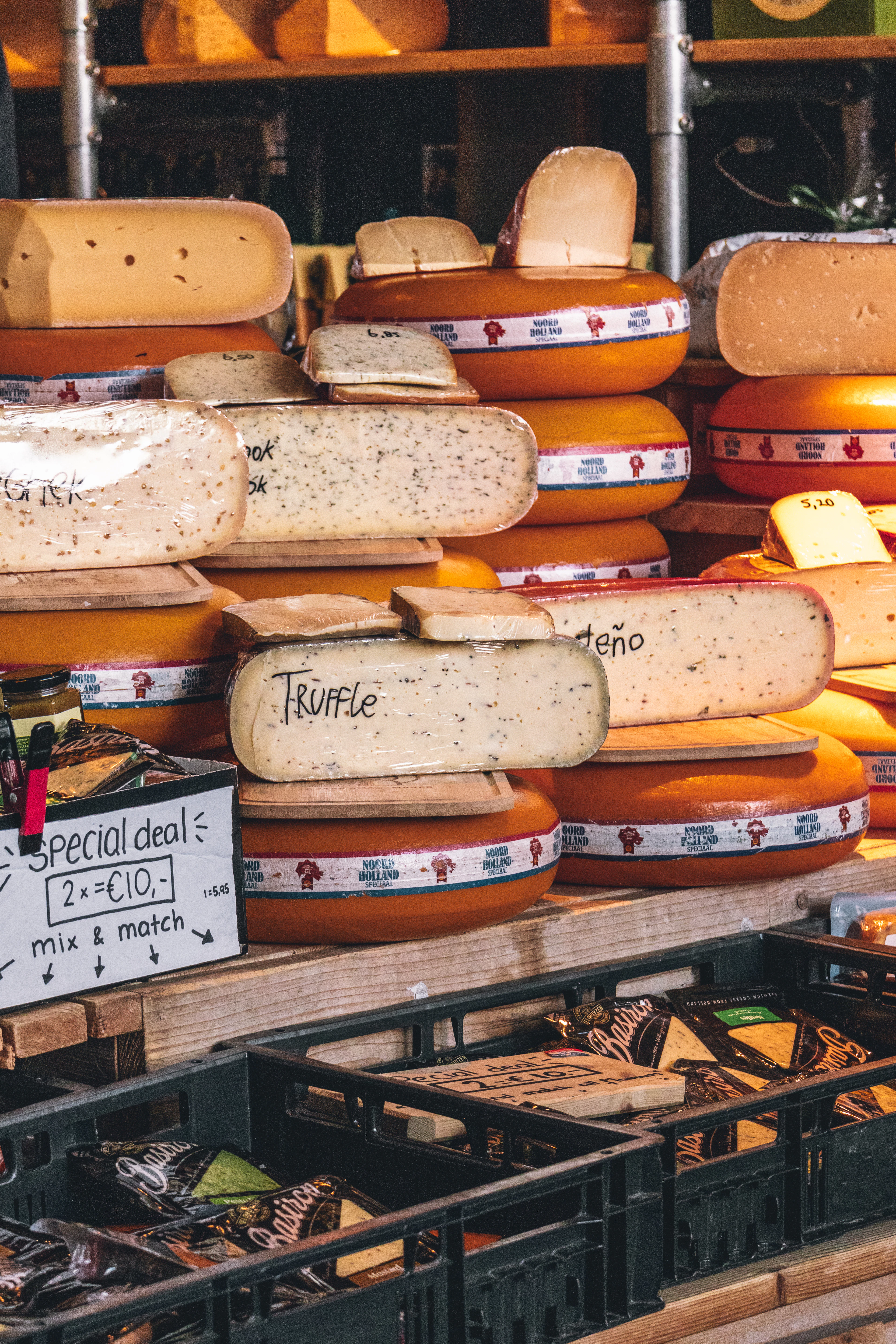 A Rotterdam market cheese stall stacked with waxed Noord-Holland gouda wheels and cut wedges labelled truffle and herb, a handwritten mix-and-match price card propped in front.
