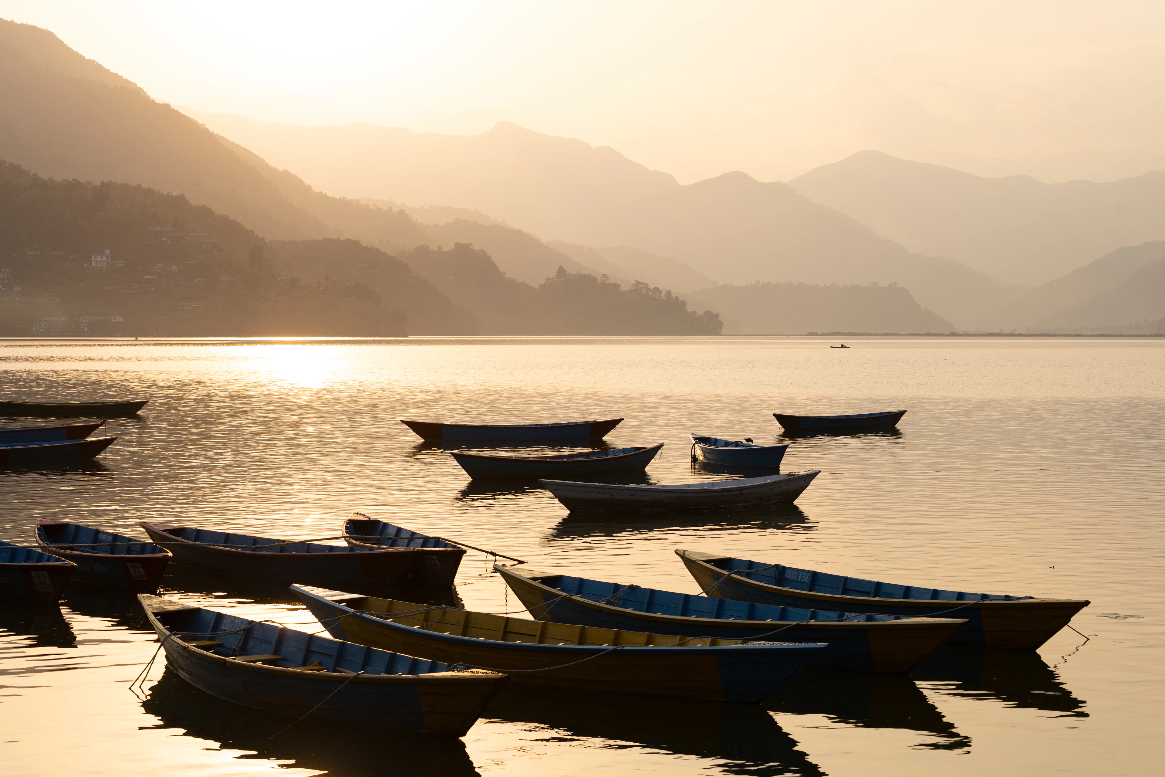 Painted wooden rowing boats drifting on a golden, mist-softened Phewa Lake at Pokhara, layered hills and a hillside village catching the low sun behind them