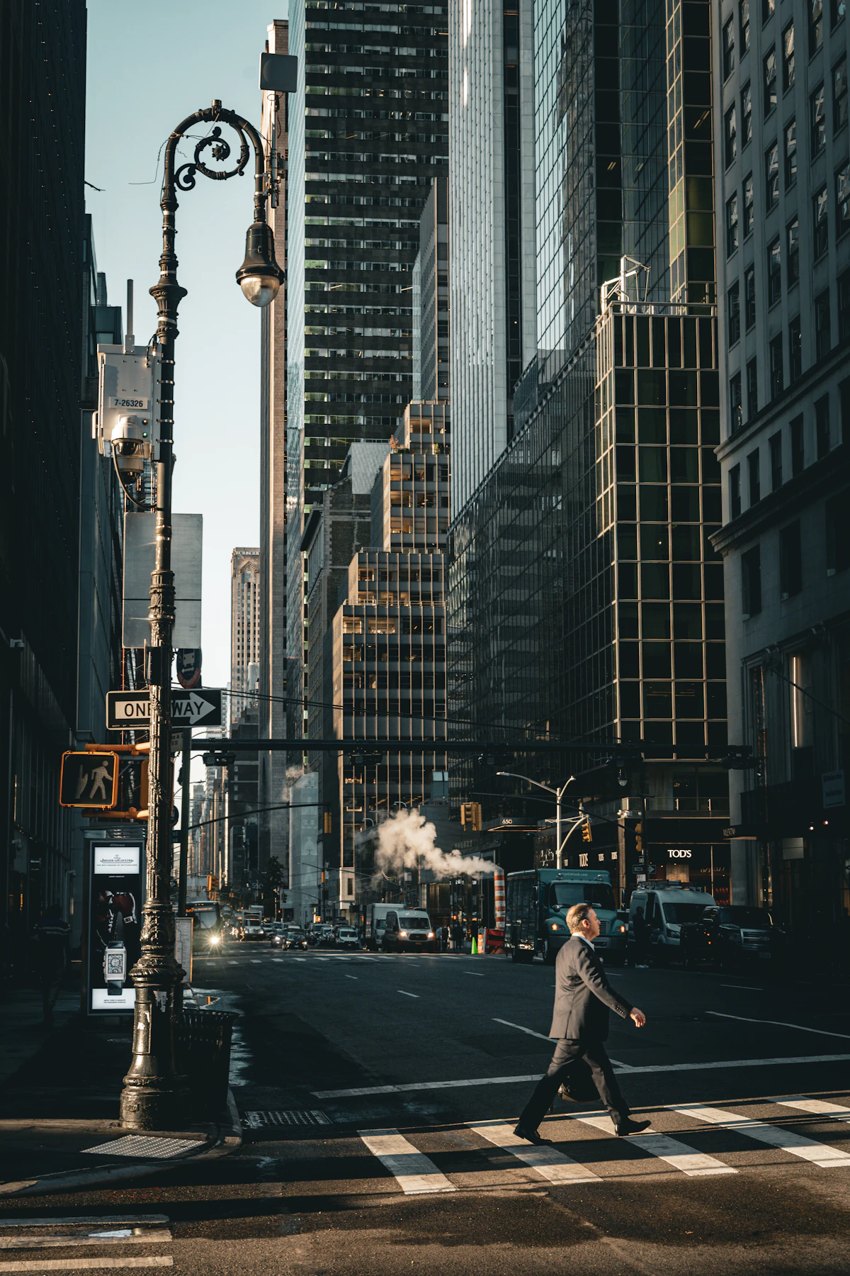 A pedestrian crossing a Manhattan avenue between tall buildings in the morning light