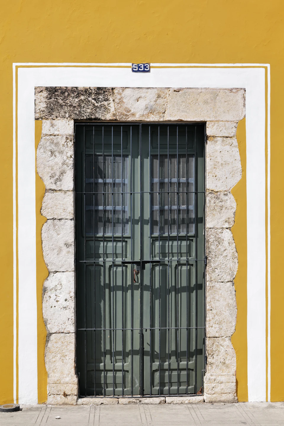 The image shows a stone doorway with two green doors, one on each side. The door is slightly open, revealing a glimpse of the interior beyond it. Above the doorway, there are white lines painted on the wall, adding an artistic touch to the scene. The colors in the image are predominantly yellow and green, creating a warm and inviting atmosphere.