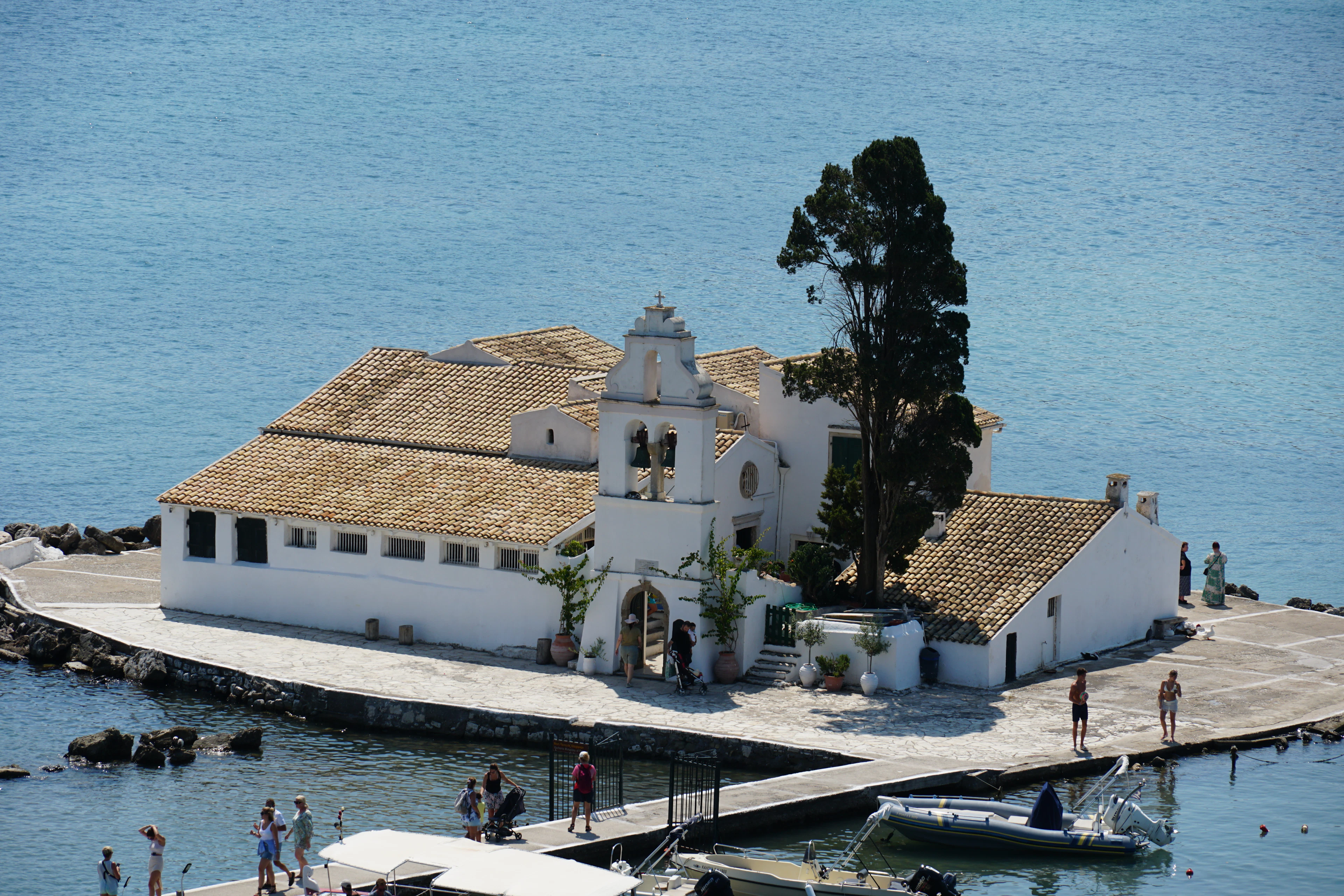 The white walls, terracotta roof and small bell tower of Vlacherna Monastery on its islet at Kanoni, Corfu, a dark cypress rising beside it and boats moored along the narrow causeway in deep blue water