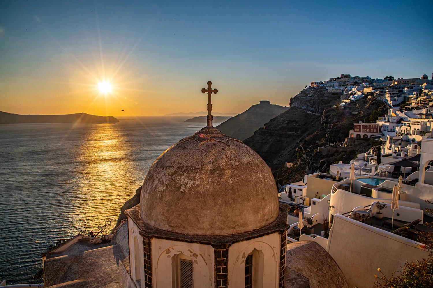 Sunrise over a Greek island village, light breaking across a domed church above the sea.
