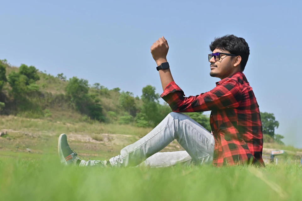 Young man sitting on hillside with arms raised, wearing plaid shirt and jeans outdoors