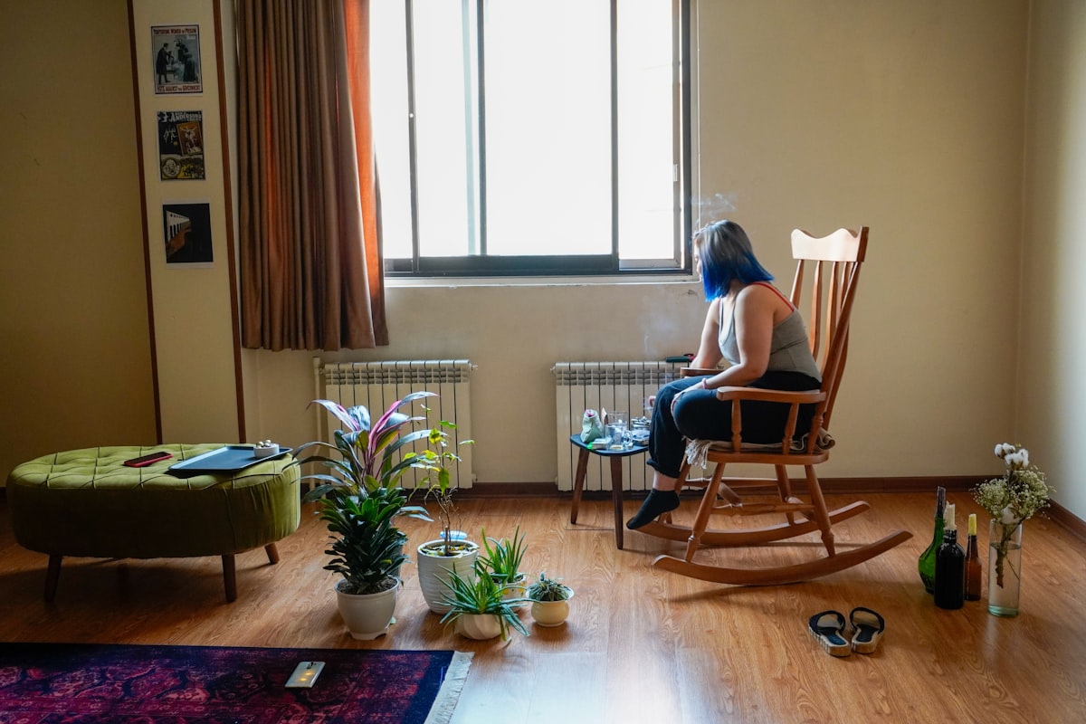 A person sits in a rocking chair in a sunlit, plant-filled room, turned toward the window, seen from behind.
