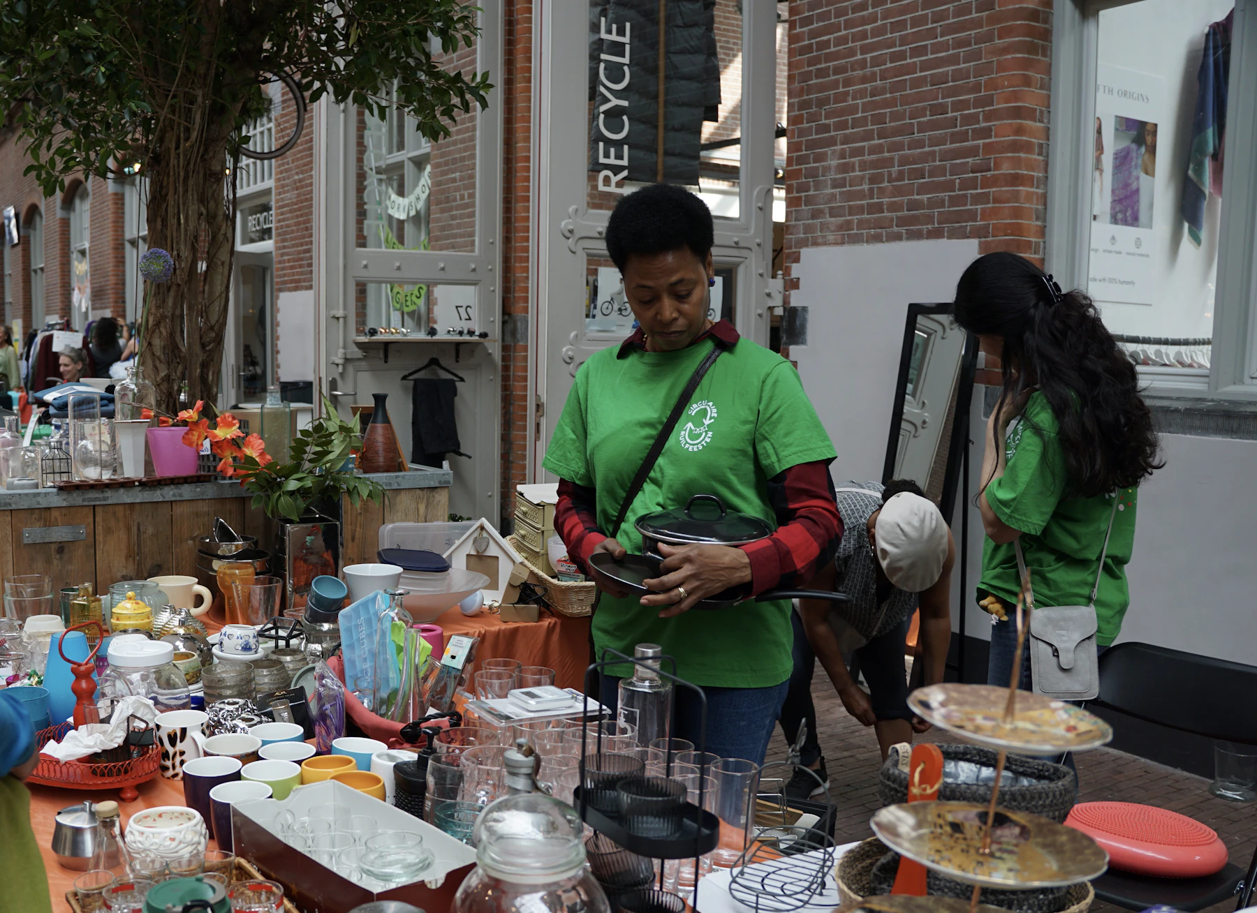 Shoppers looking over an outdoor yard sale table crowded with glassware and dishes