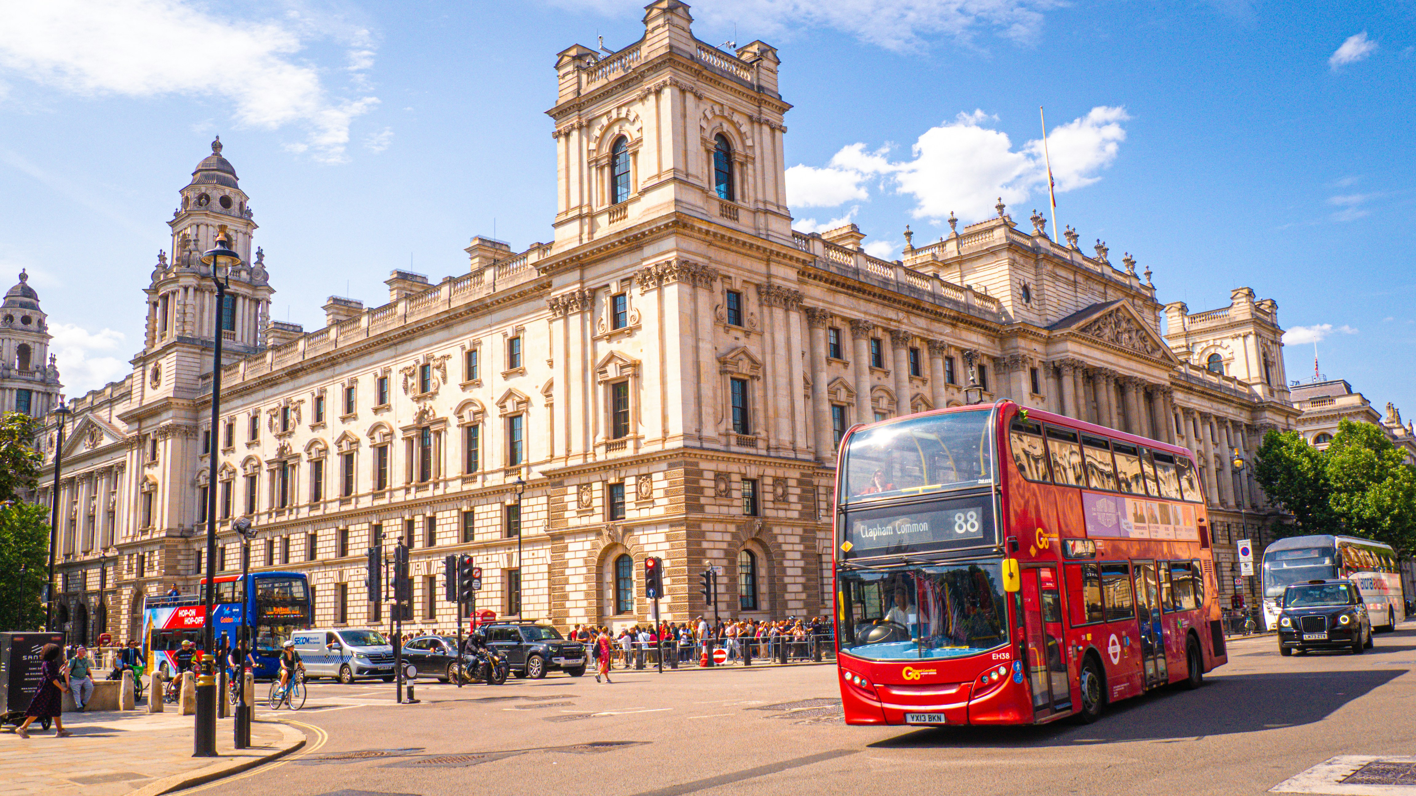 A red double-decker bus drives past a historic building.
