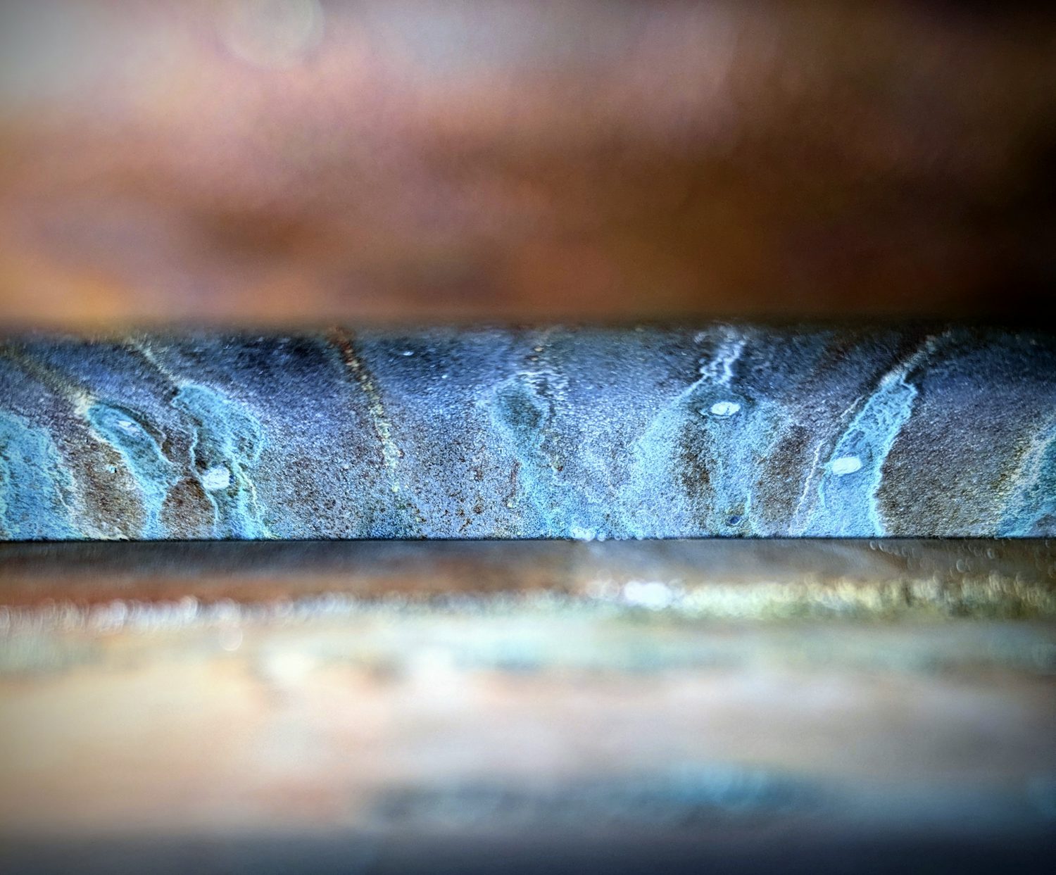 A close-up of the blue-y green rust within my water heater. It can only be seen through a small horizontal slot in the centre, with the rest of the shot showing the out-of-focus exterior of the heater.