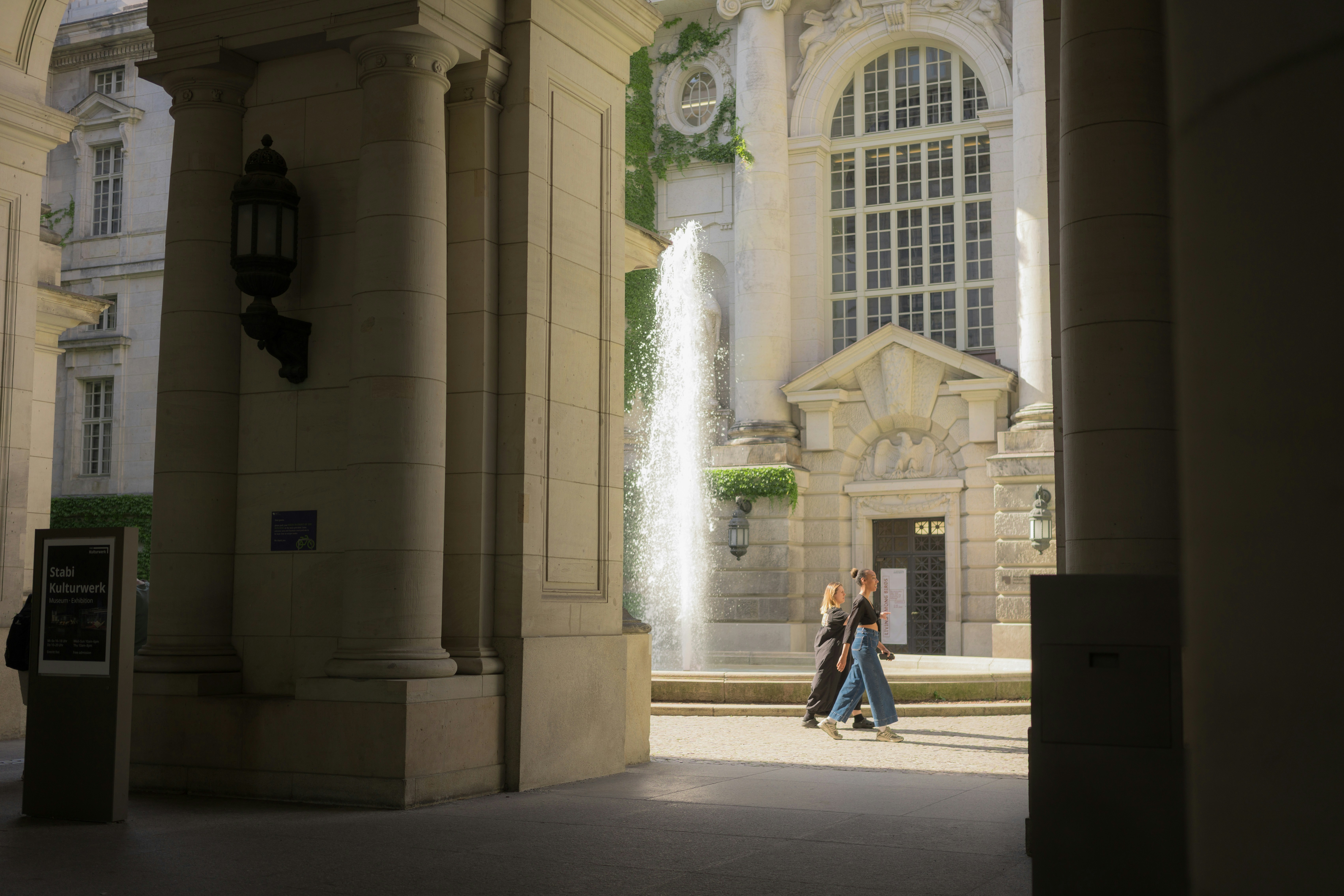 Two people walk past a fountain in an archway.