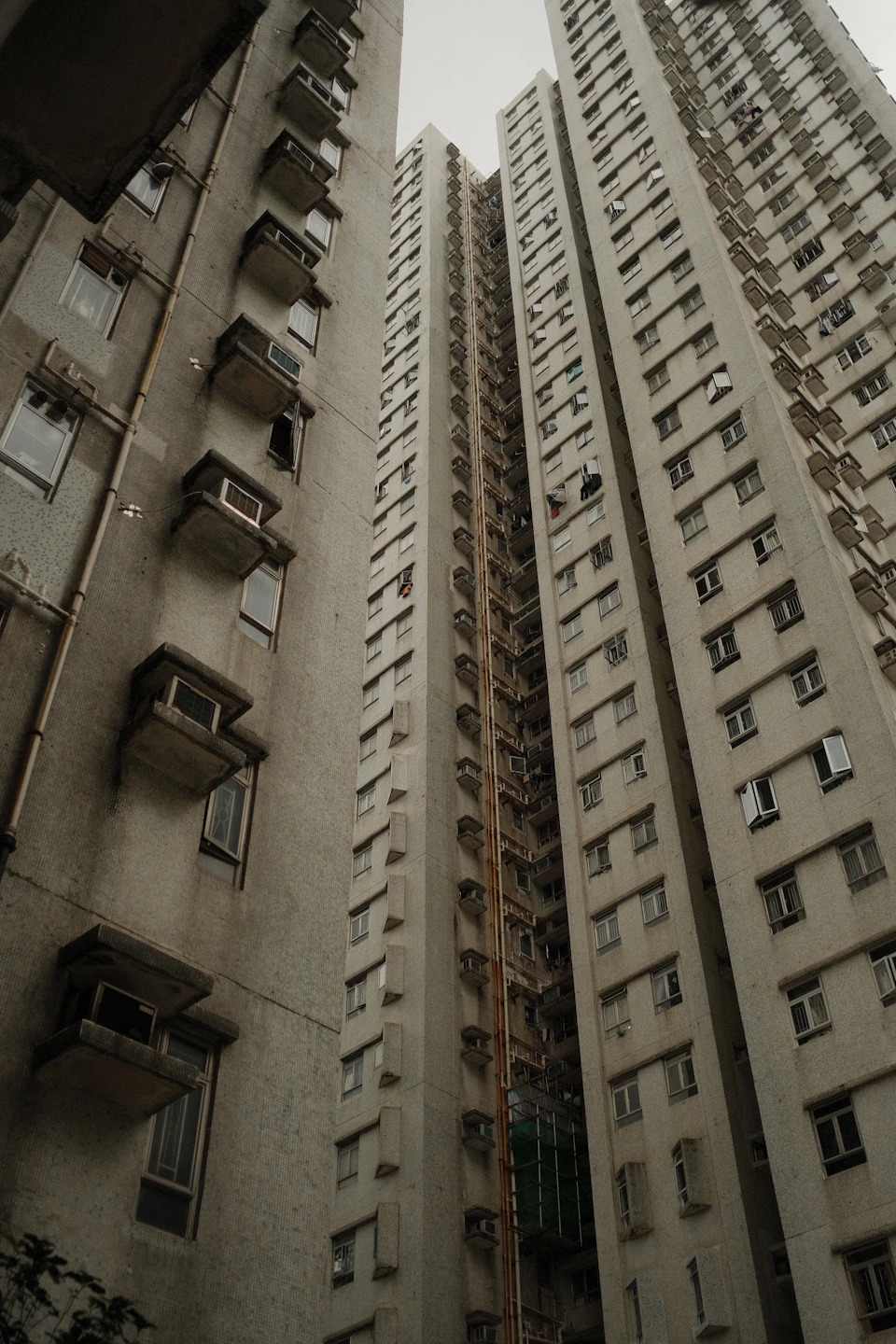 The image depicts a tall, gray apartment building with multiple levels and balconies on each floor. The building is situated between two other structures, creating an urban landscape. The perspective of the photo is from below, looking up at the imposing height of the structure.