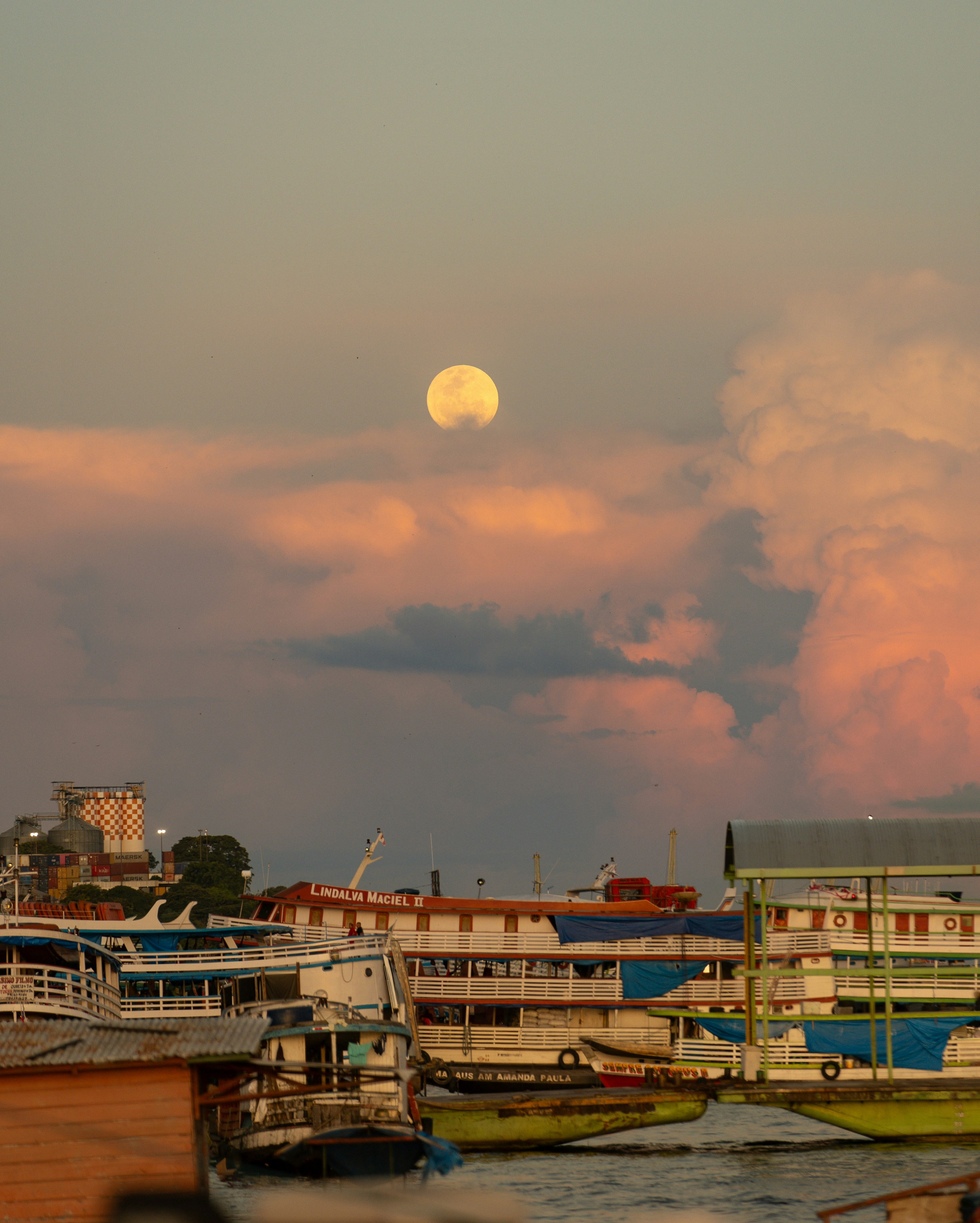 A lua sobe sobre barcos e nuvens coloridas do pôr do sol.