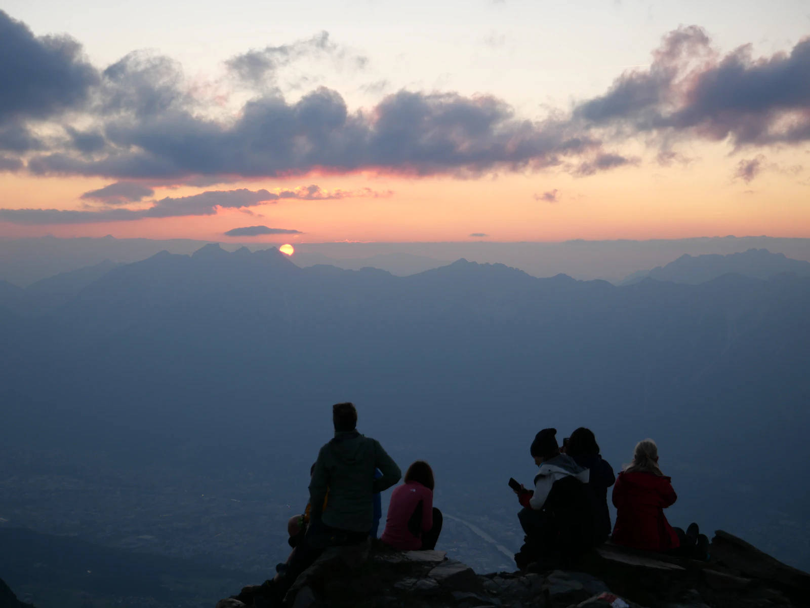 Photograph of People sitting beside an Austrian mountain hut while the sun lowers beyond distant ridges.