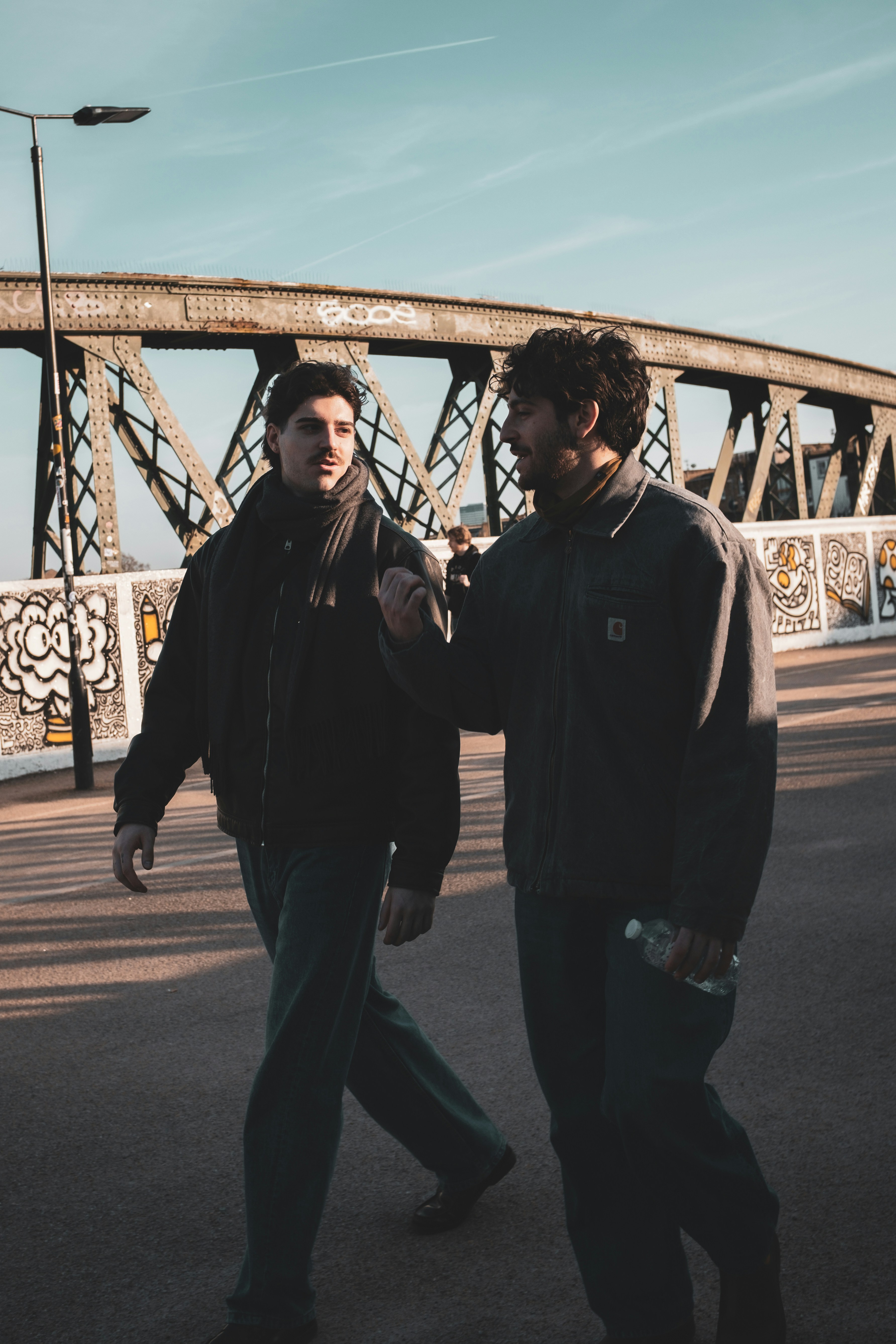 Two men walk and talk on a bridge.