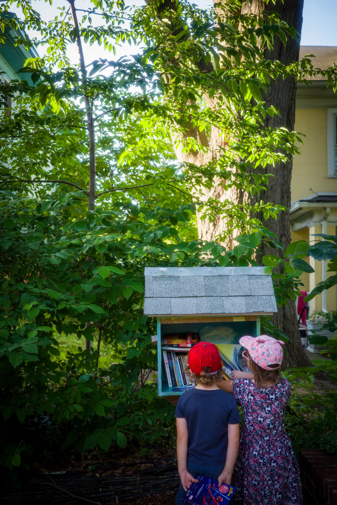 Two kids browsing books in a little free library in a leafy yard
