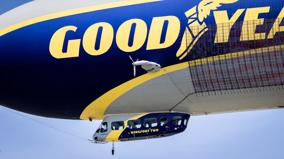 The image shows a large blue and yellow airplane soaring through the sky, with its nose pointed towards the bottom right corner. The airplane is adorned with the words "Good Year" on its side, indicating that it belongs to the Goodyear company. Below the airplane, there's a smaller airplane painted in shades of blue and white, positioned at an angle relative to the larger aircraft.
