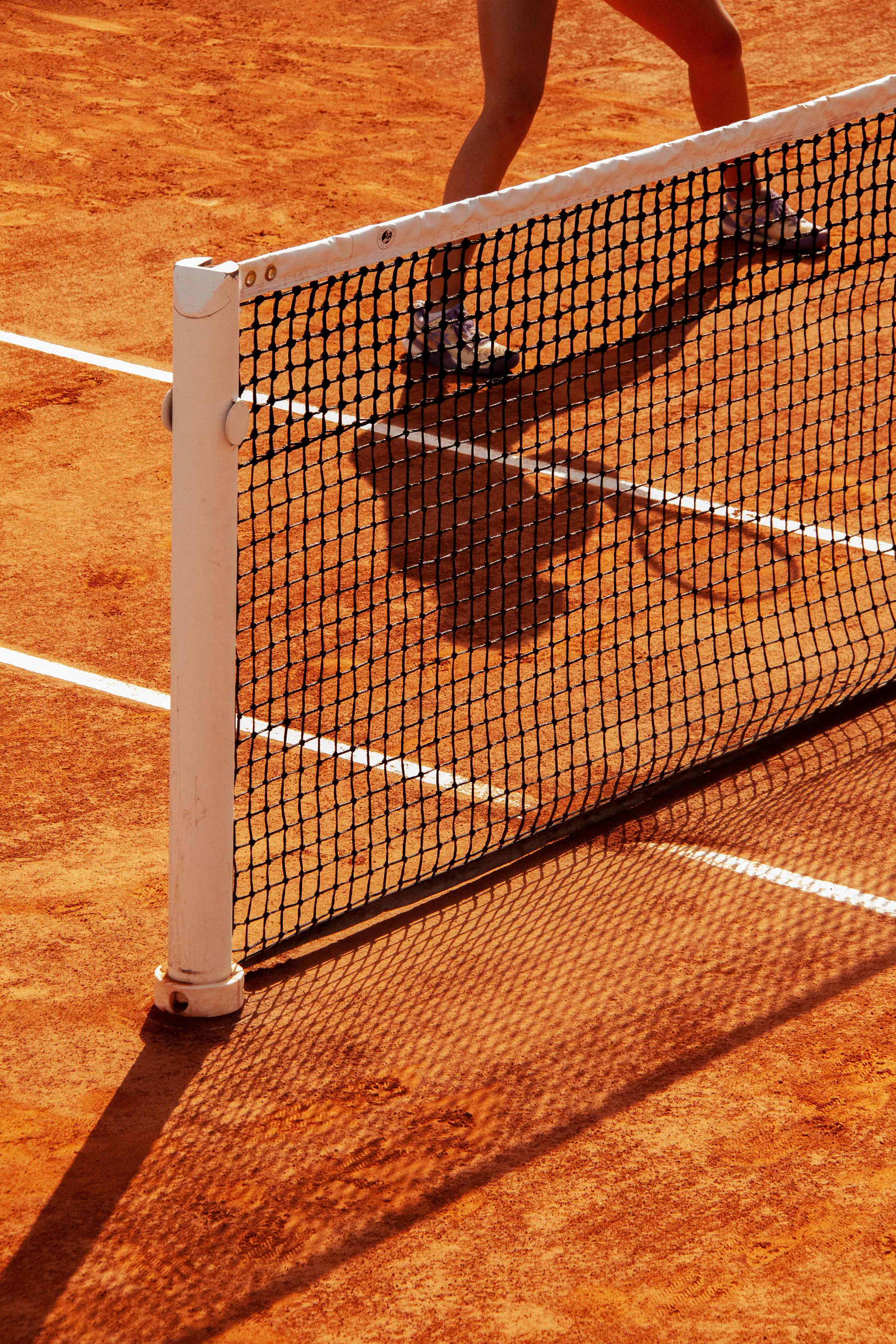 A tennis net post and net over red clay, the shadow of a player behind it