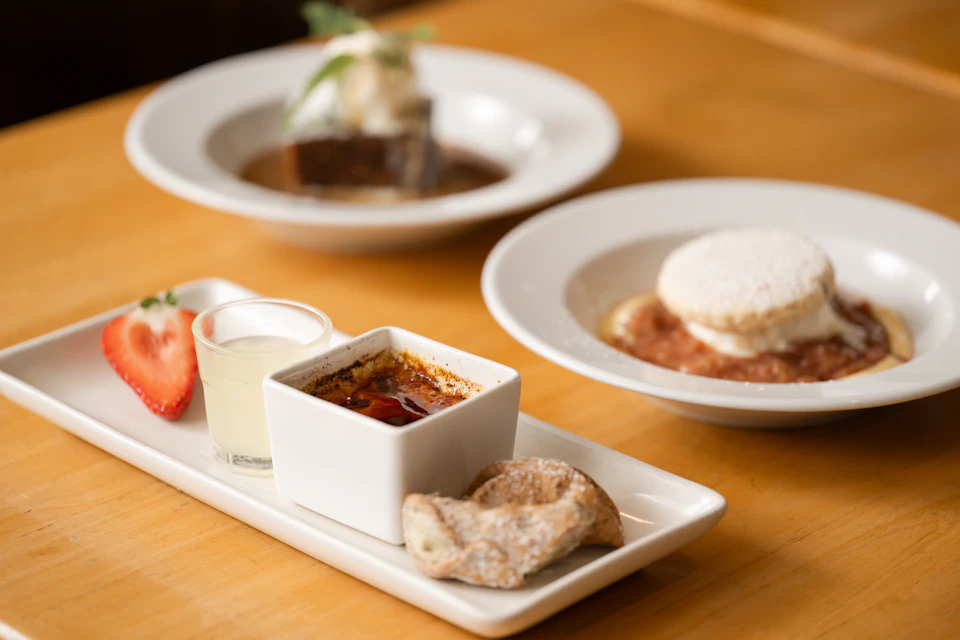 The image shows a wooden table with three plates of food arranged on it, including a small white bowl filled with a dessert and two larger bowls containing different dishes. The desserts are topped with strawberries, adding a pop of color to the scene. In addition to the main course, there is also a small cup of tea placed next to one of the bowls.

The table itself has a warm brown hue, providing a cozy atmosphere for the meal.