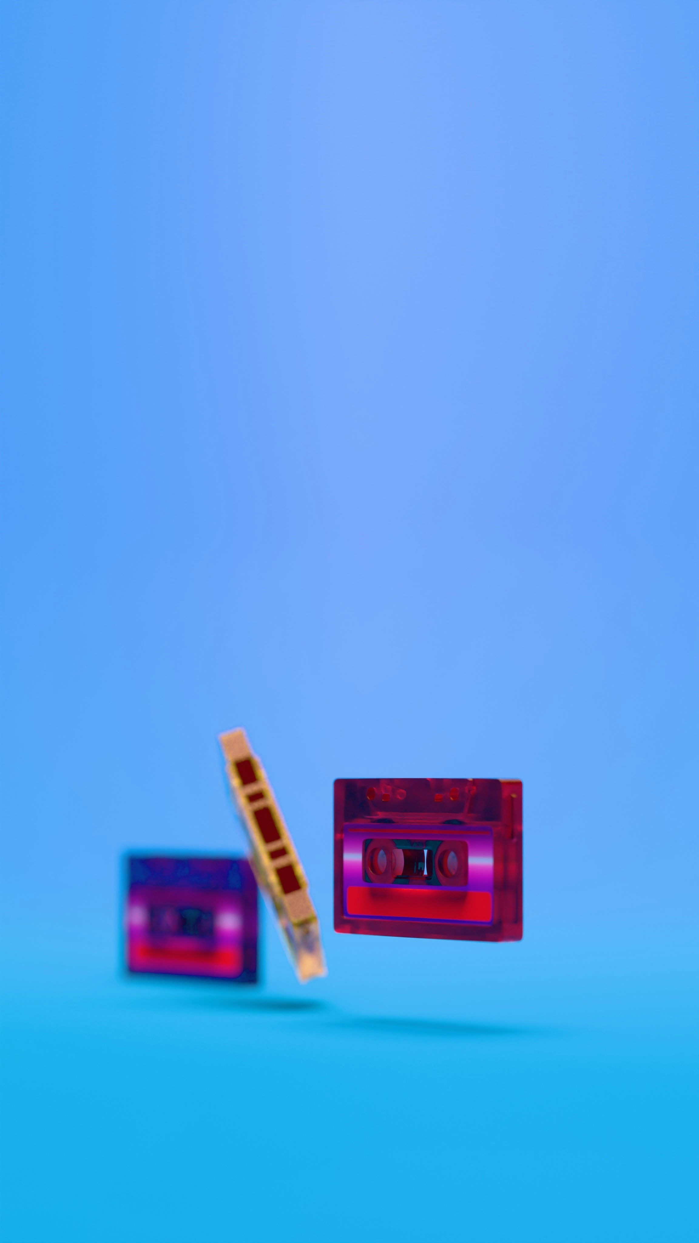 Retro audio cassette tapes rendered in glossy translucent plastic appear suspended or aligned in space over a seamless blue backdrop, highlighting form, vibrant color and nostalgic technology.
