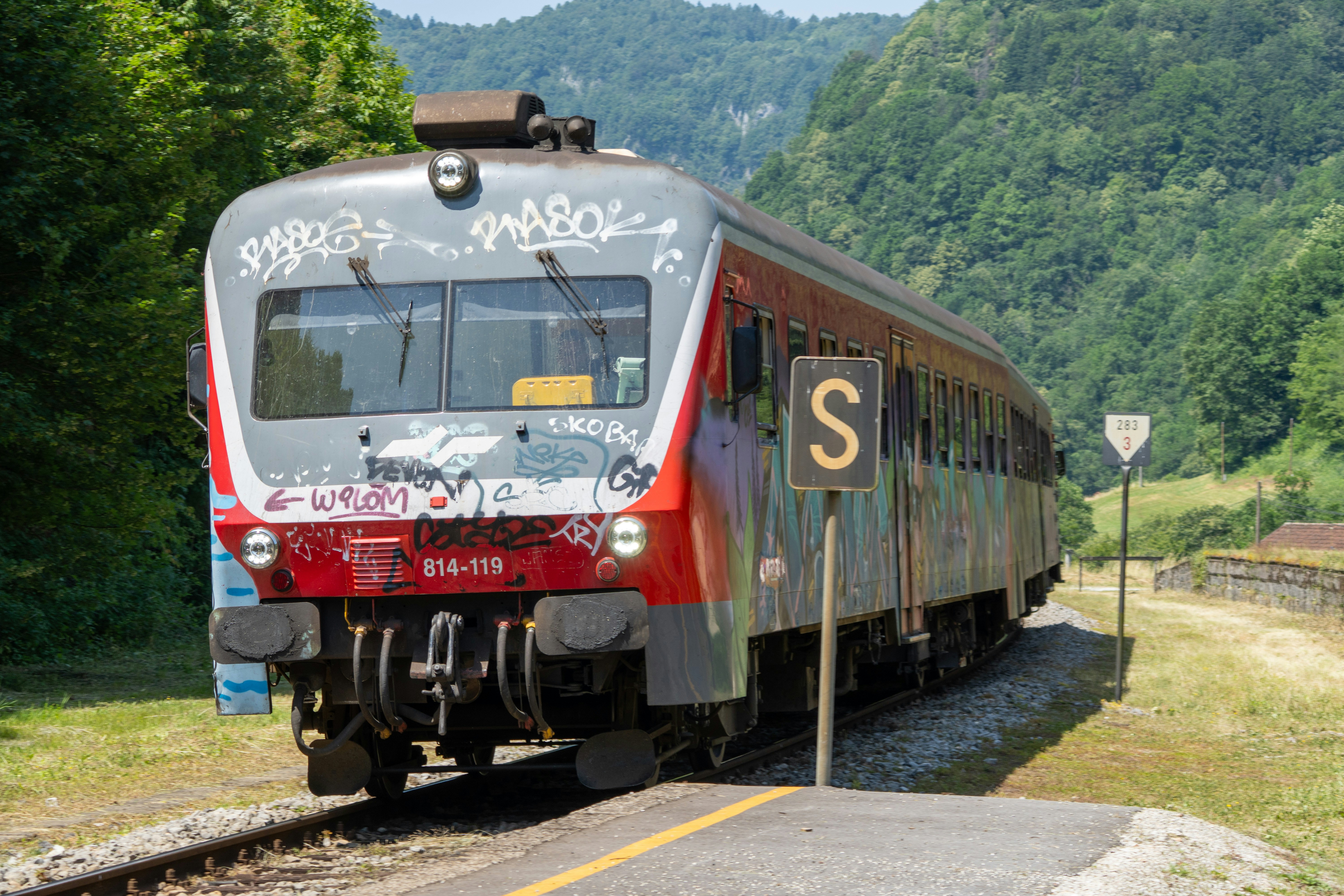 A train travels along tracks surrounded by nature., A graffiti-covered red train pulls into a rural Slovenian station, curving along tracks framed by lush green hills. The contrast between urban street art and natural surroundings creates a compelling visual of movement, culture, and landscape.