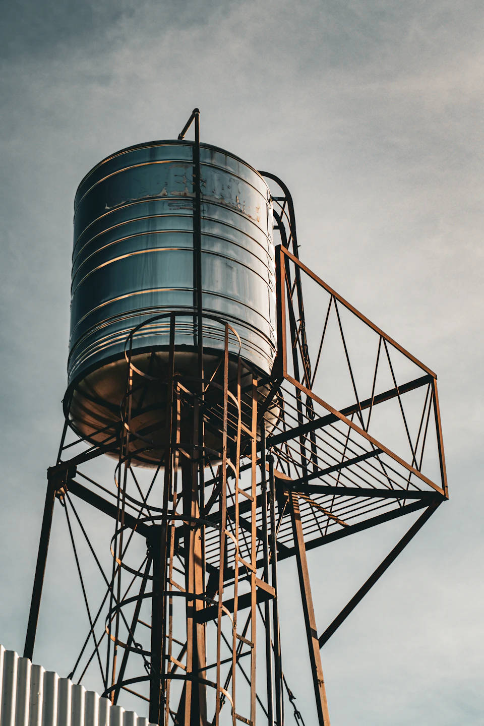 The image depicts a large, cylindrical water tank with a silver exterior and a black metal frame at its base. The tank is situated on a tall, rusted metal pole that stands against an overcast sky. A ladder is attached to the pole, leading up to the top of the tank.