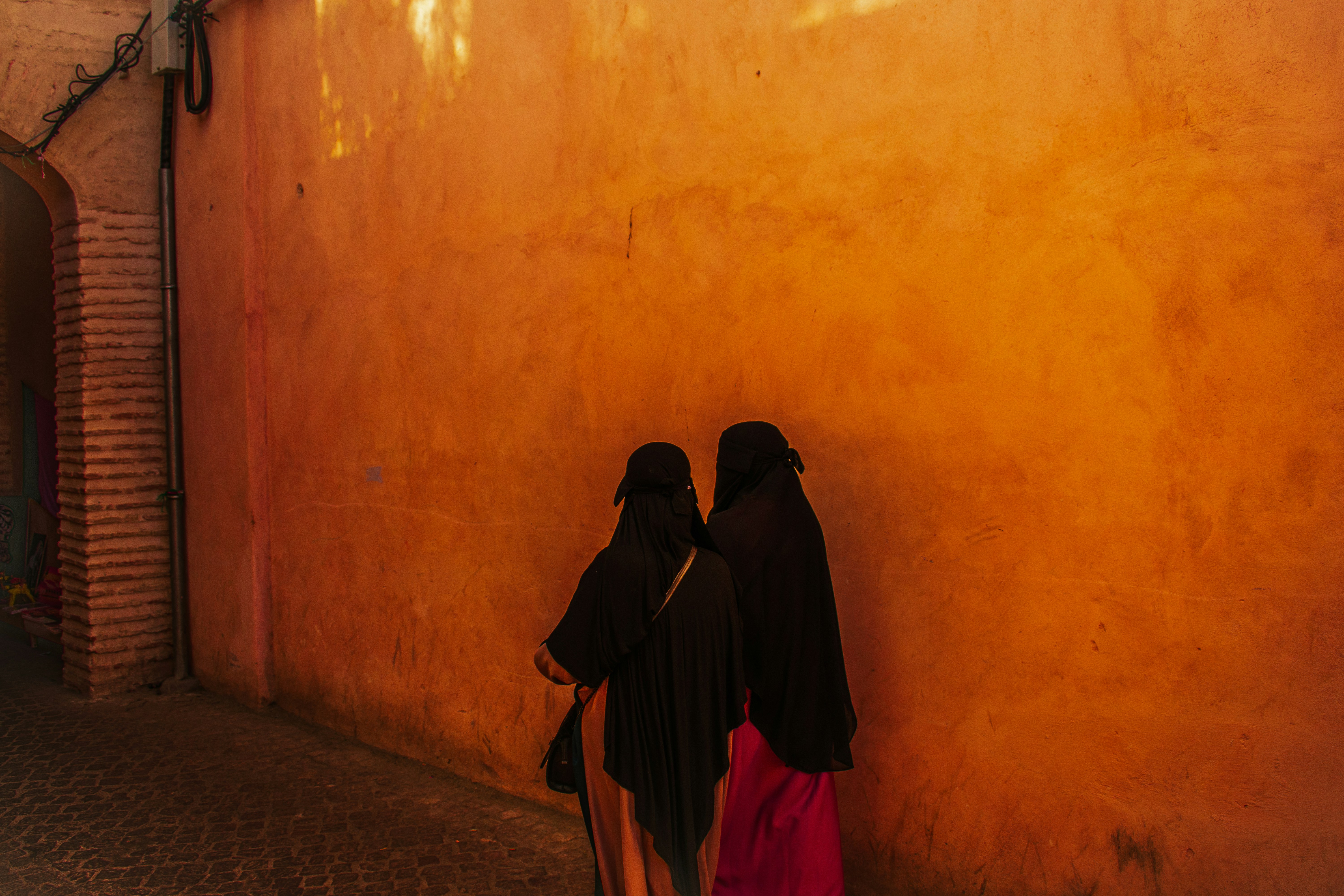 Due donne vestite di nero camminano lungo un vicolo illuminato dal sole.