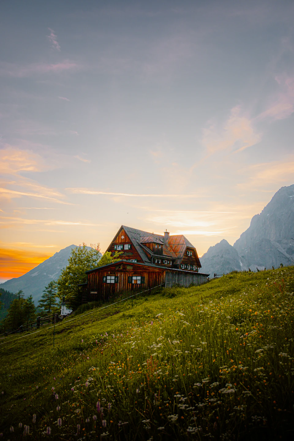 Rustic wooden cabin on grassy hill surrounded by wildflowers and mountains under clear blue sky