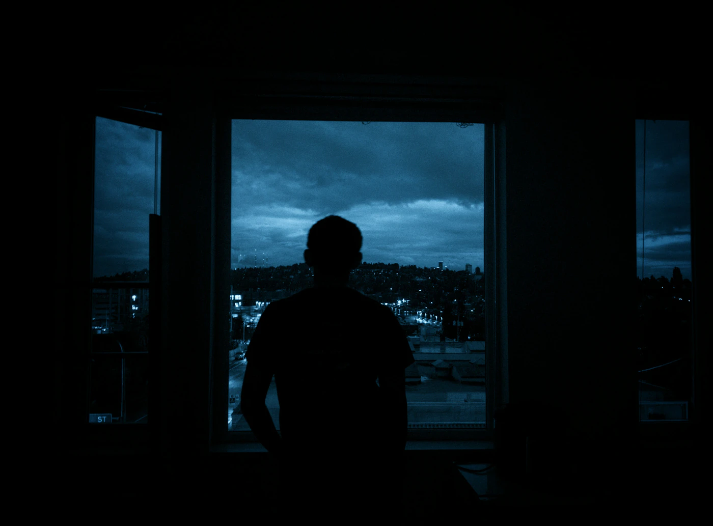 A silhouetted figure looking out a tall window at a moody city skyline at night, an image of relocation loneliness