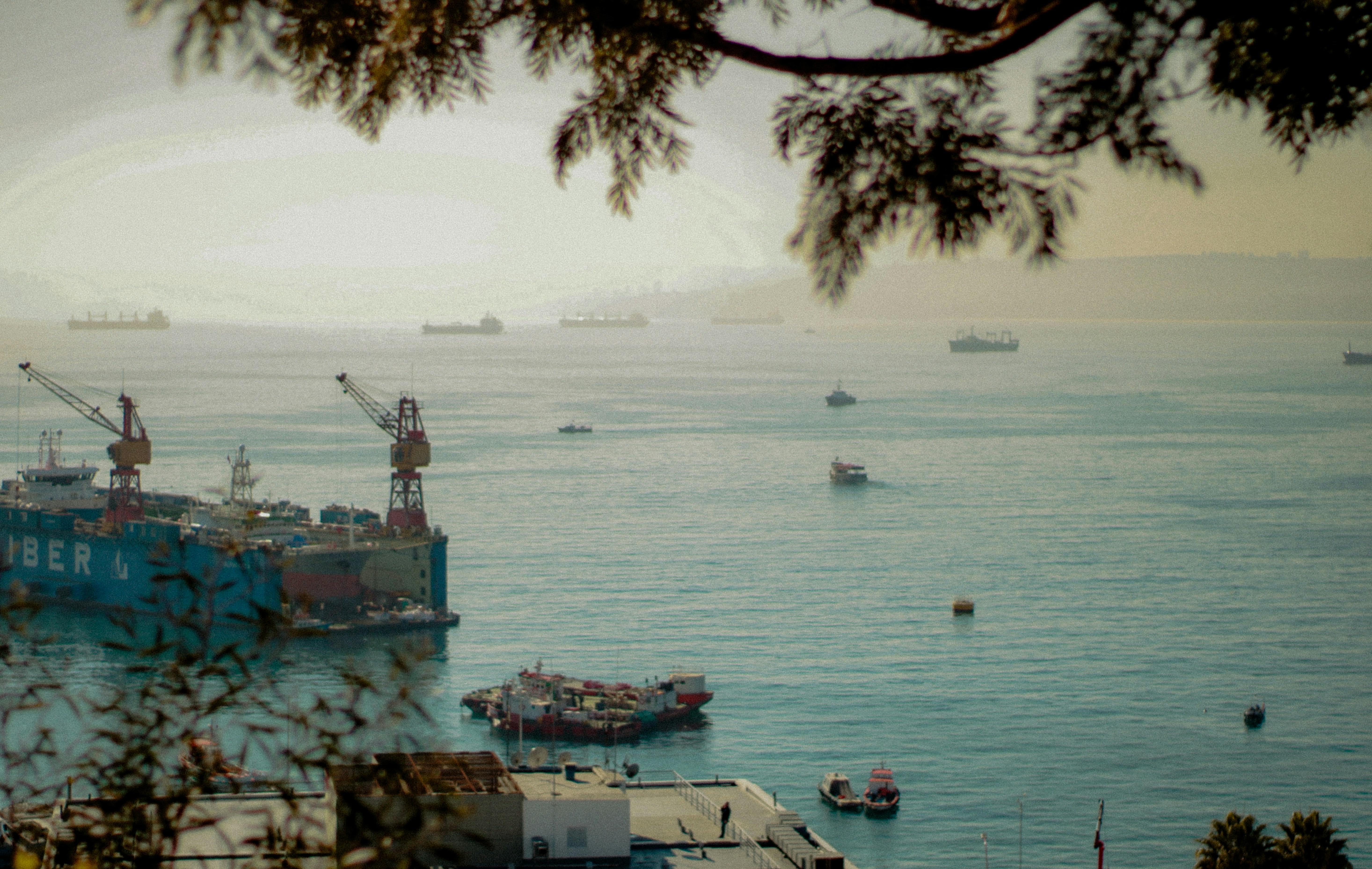 Boats and cranes fill a hazy seascape.