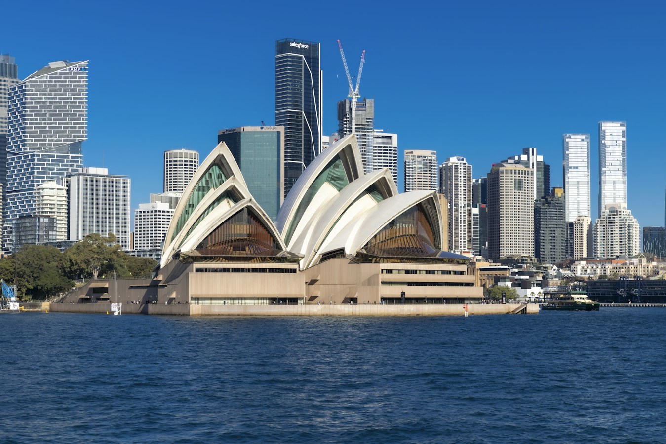 The Sydney Opera House with its white sail roofs sits on the harbour foreshore in front of a cluster of glass office towers under a clear deep-blue