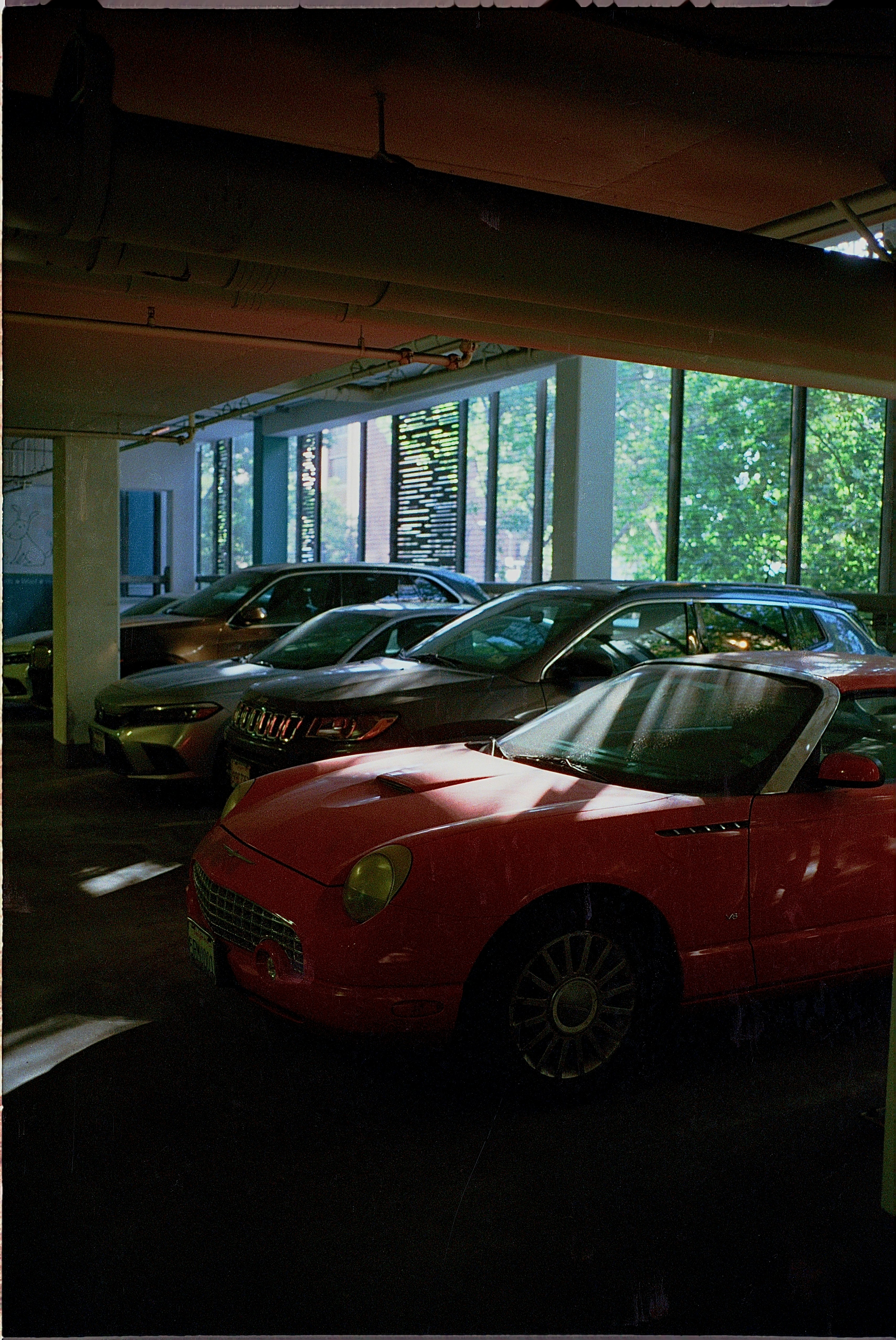 A vibrant red convertible parked among a lineup of modern vehicles in an urban garage, illuminated by natural light streaming through large windows.
