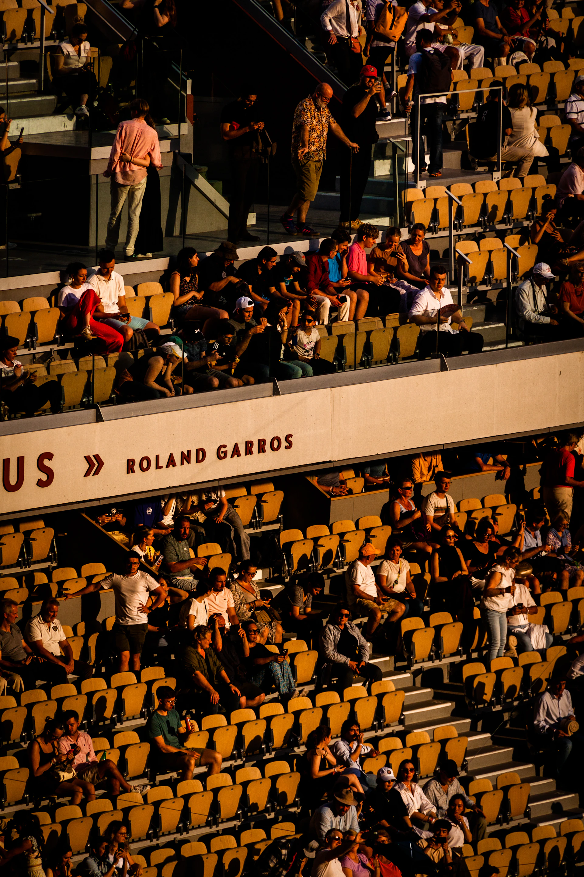 Трибуны корта Филиппа Шатрие с вывеской ROLAND GARROS, заполненные зрителями в лучах закатного солнца