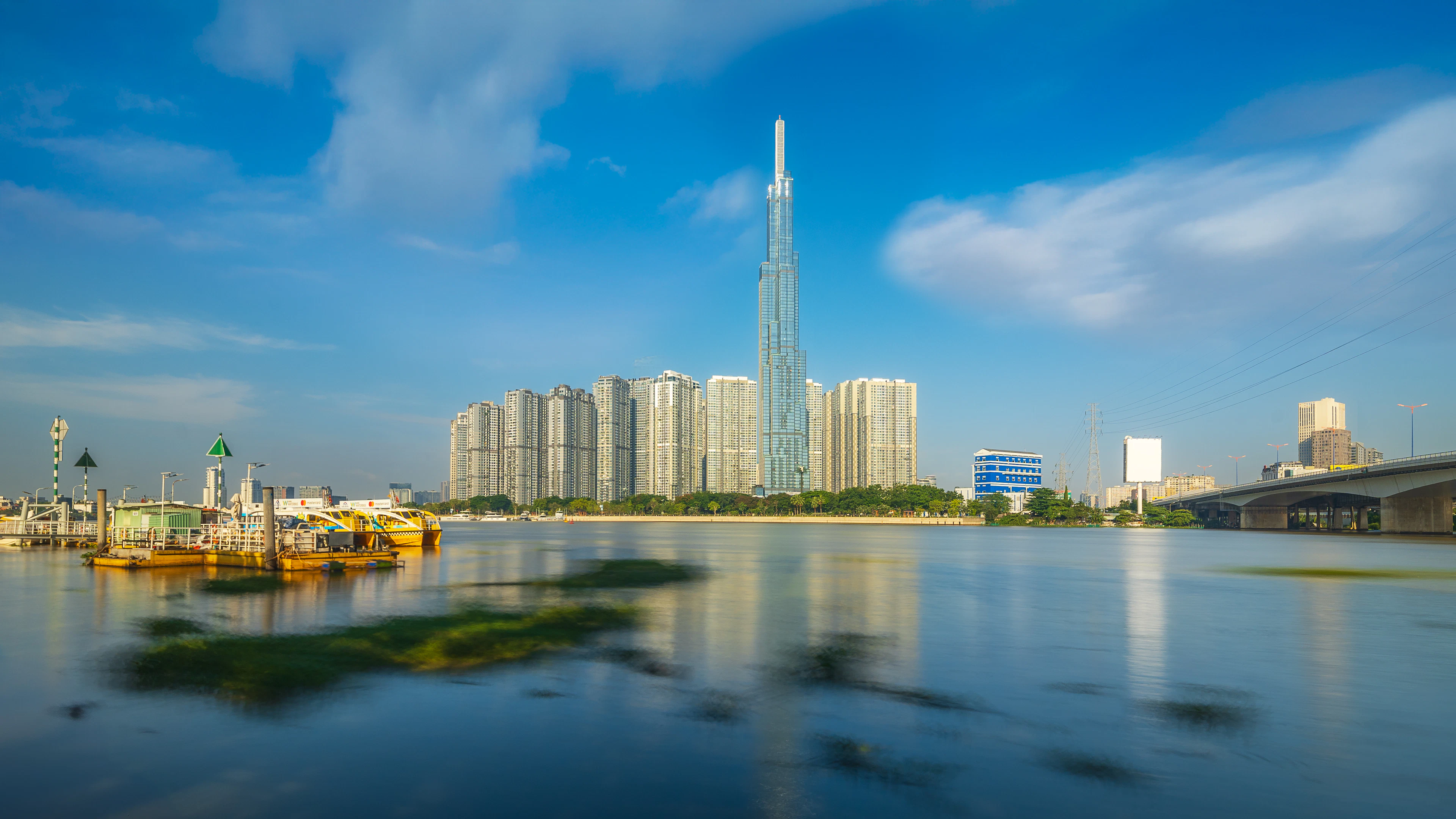 The Landmark 81 tower and the riverside high-rises of Ho Chi Minh City rising above the Saigon River, mirrored in still water under a bright blue sky streaked with white cloud.