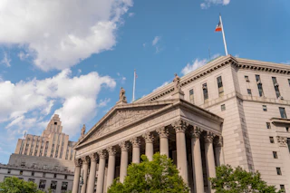 A limestone courthouse with a columned portico, statues on the pediment, and a flag against a blue sky.