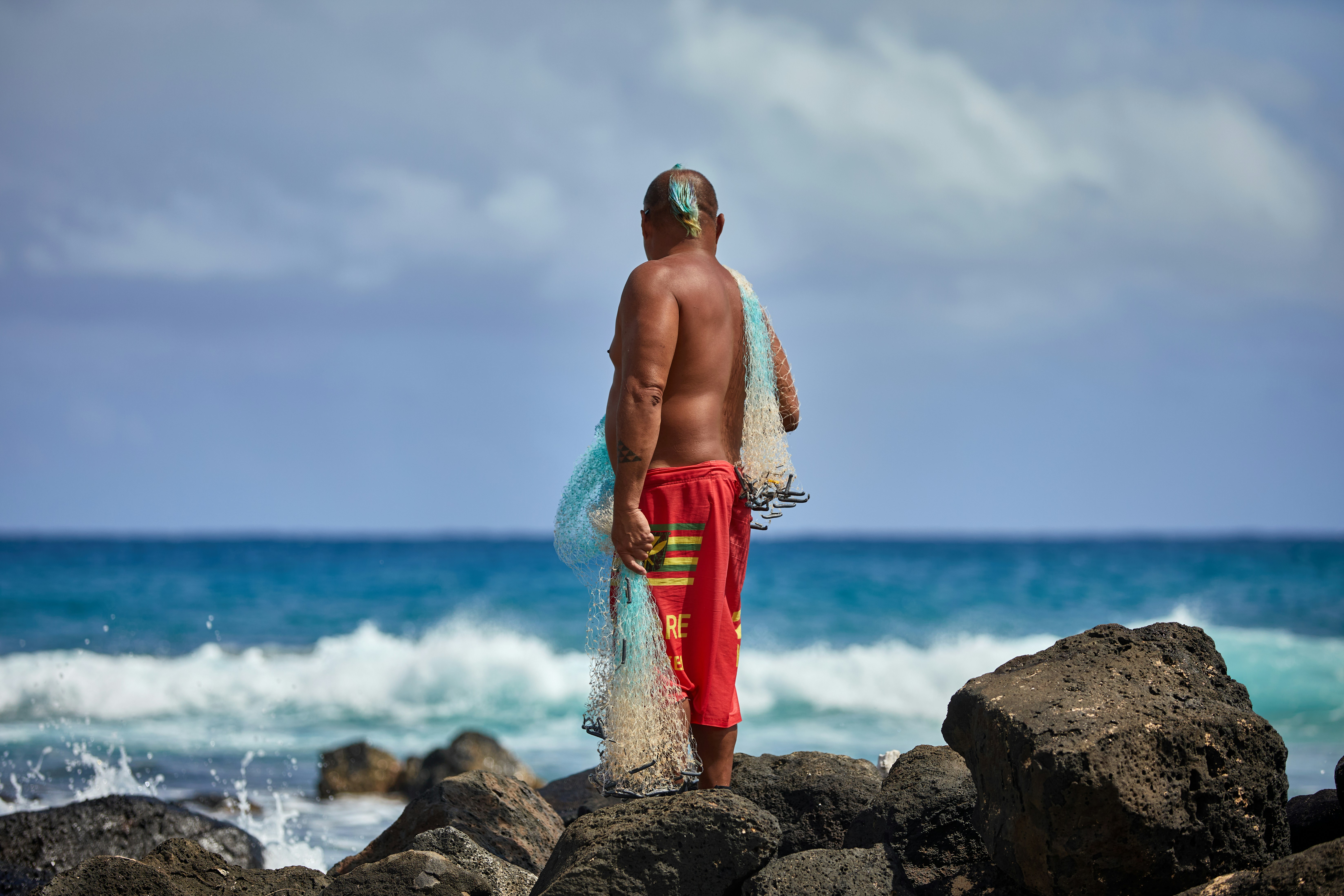 A fisherman stands on Hawaiian coastal rocks with his net, representing the active outdoor lifestyle that helps Hawaiians live nearly 80 years.